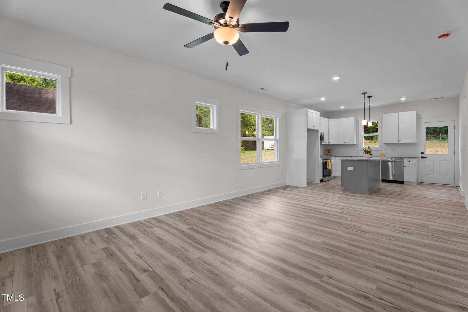 Open-plan living space with light wood flooring, white walls, and a ceiling fan; kitchen area visible in the background with white cabinets, stainless steel appliances, and an island. Three windows and a back door provide natural light.