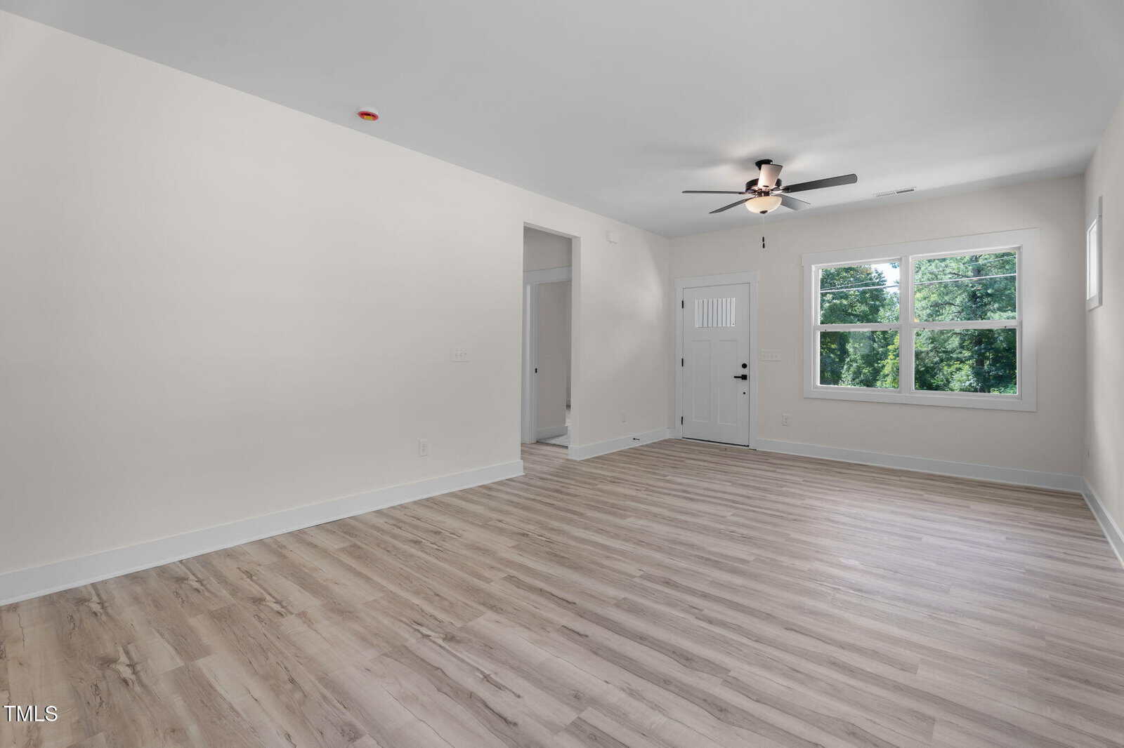Empty room with light wood flooring, white walls, a ceiling fan, a window showing greenery outside, and a white door. An open doorway leads to another area. Room is well-lit with natural light.