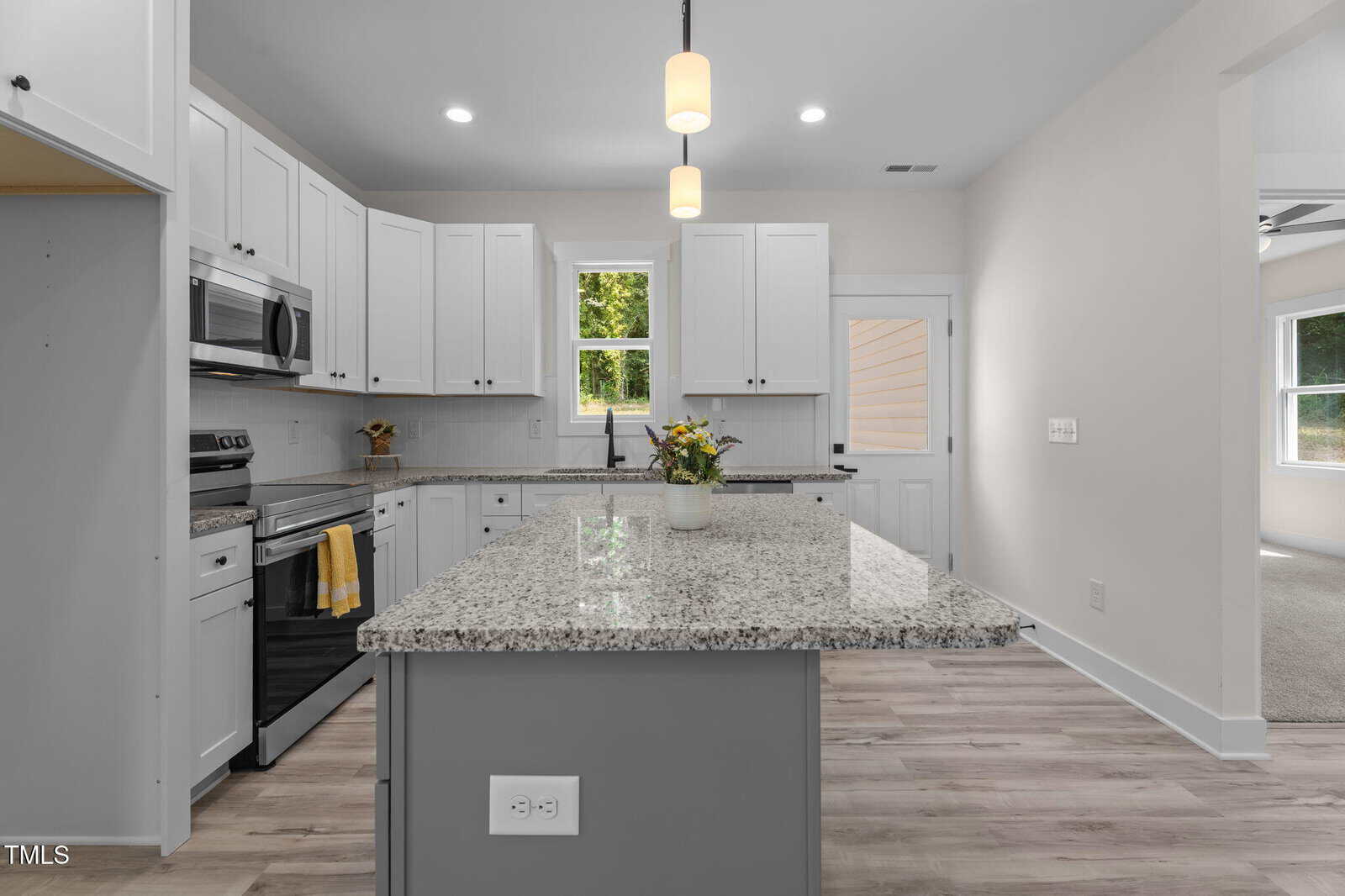 Modern kitchen with white cabinets, granite countertops, stainless steel appliances, and a center island with a potted plant. Two pendant lights hang above the island. A window and door are in the background.