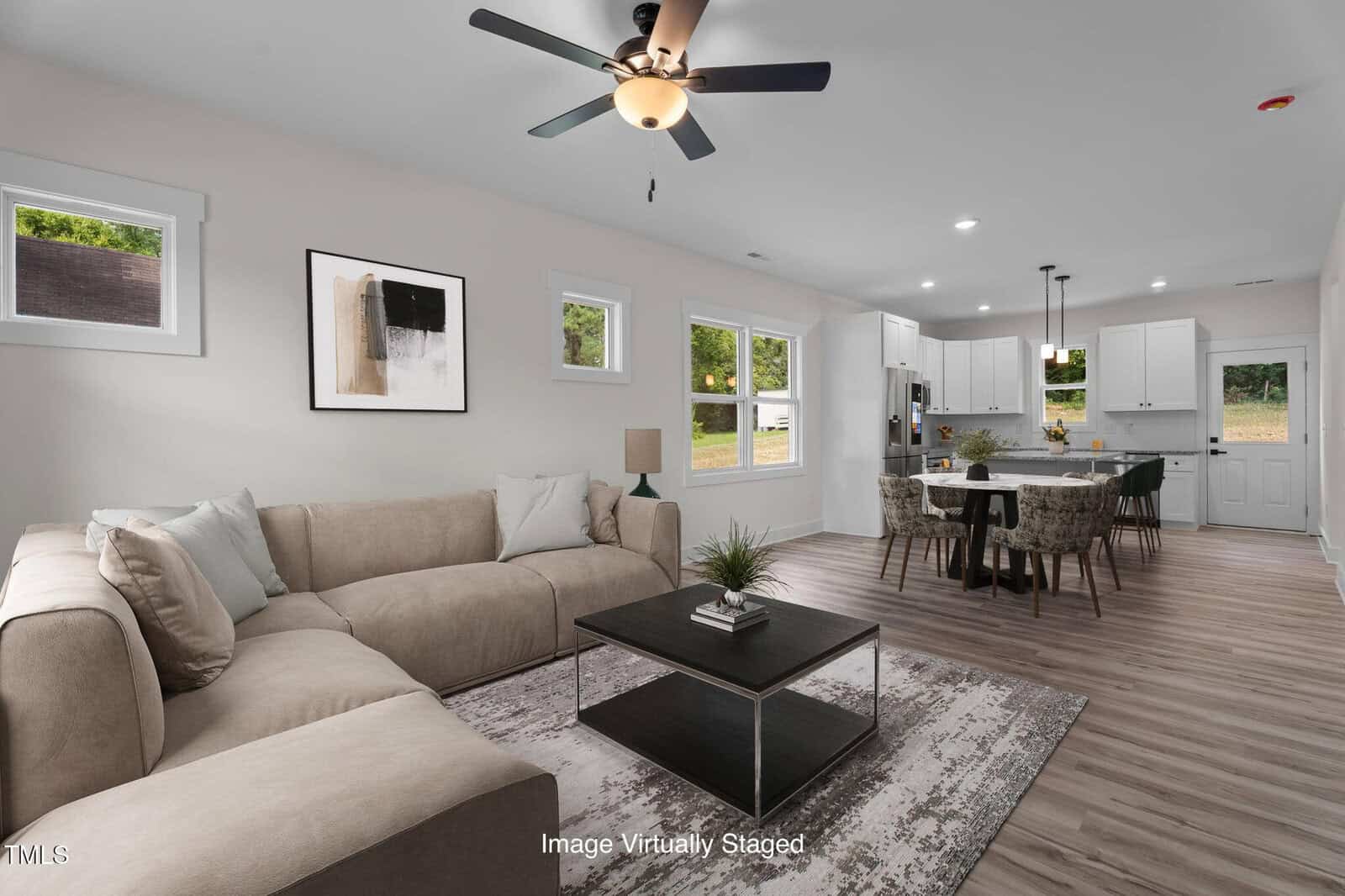 A modern, open-concept living and dining area with a beige sectional sofa, coffee table, and rug. The dining space has a table and chairs. The kitchen features white cabinets and pendant lighting. The room is virtually staged.