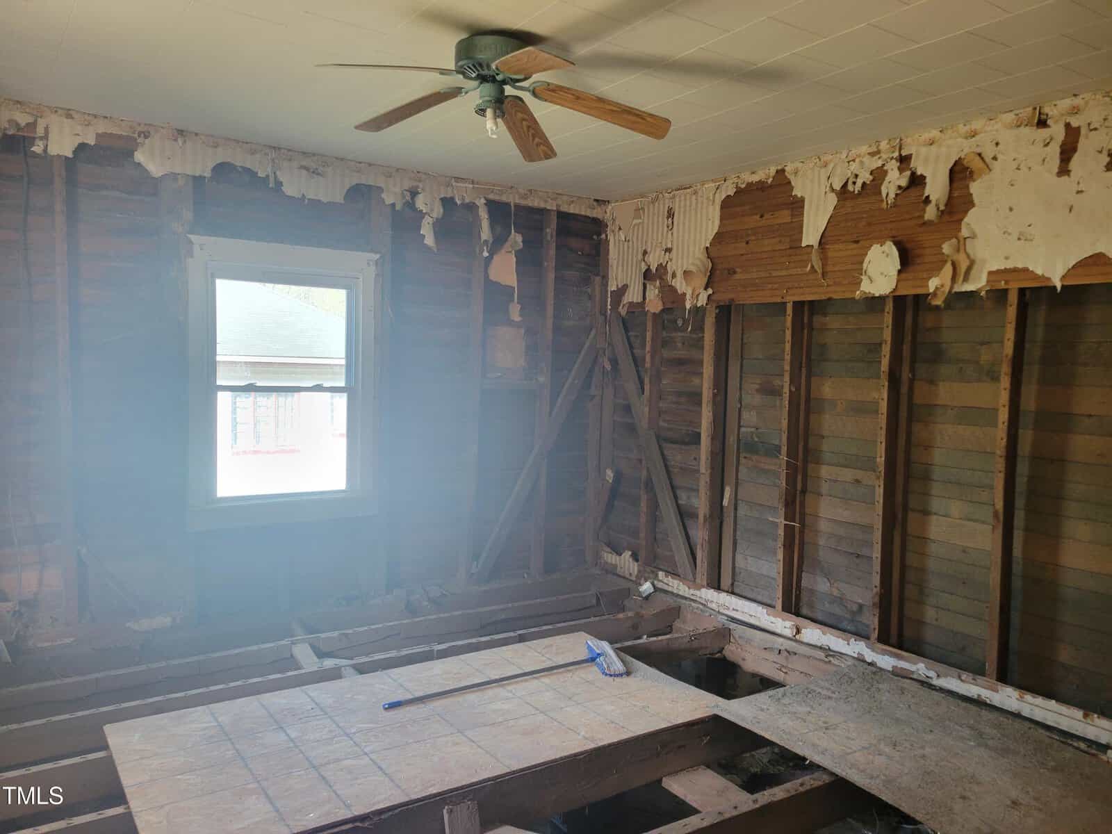 A partially demolished room with exposed wooden wall studs and a missing floor. Peeling wallpaper and an intact ceiling with a fan are visible. Sunlight enters through a window on the left, and debris is scattered on a large wooden board.