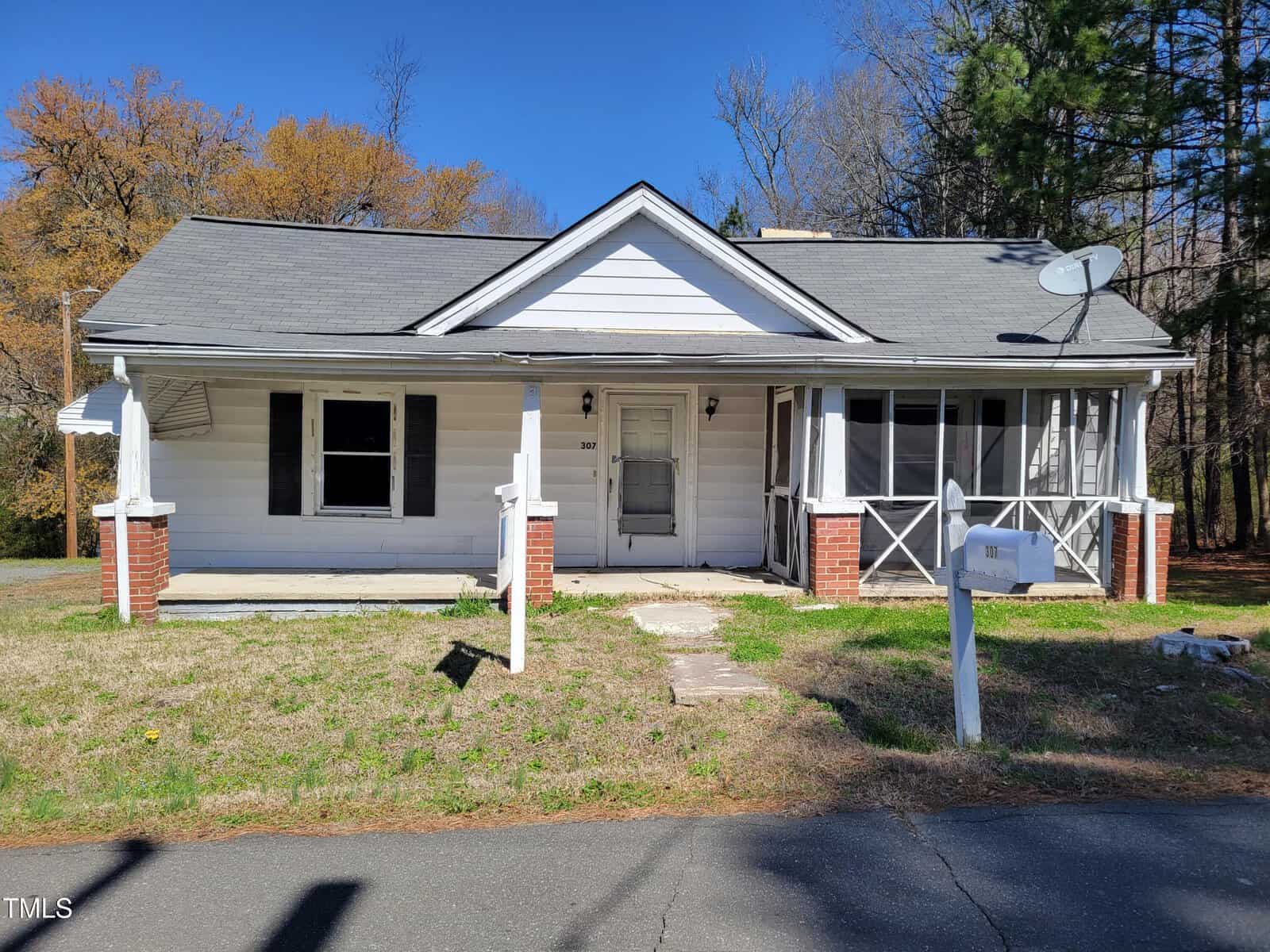 Located at 307 Watson Street in the cozy town of Creedmoor, this single-story white house features a gray roof and welcoming front porch adorned with screened windows, despite a few missing panels. A satellite dish sits above, while the mailbox stands guard by the driveway amidst sparse grass and distant trees.
