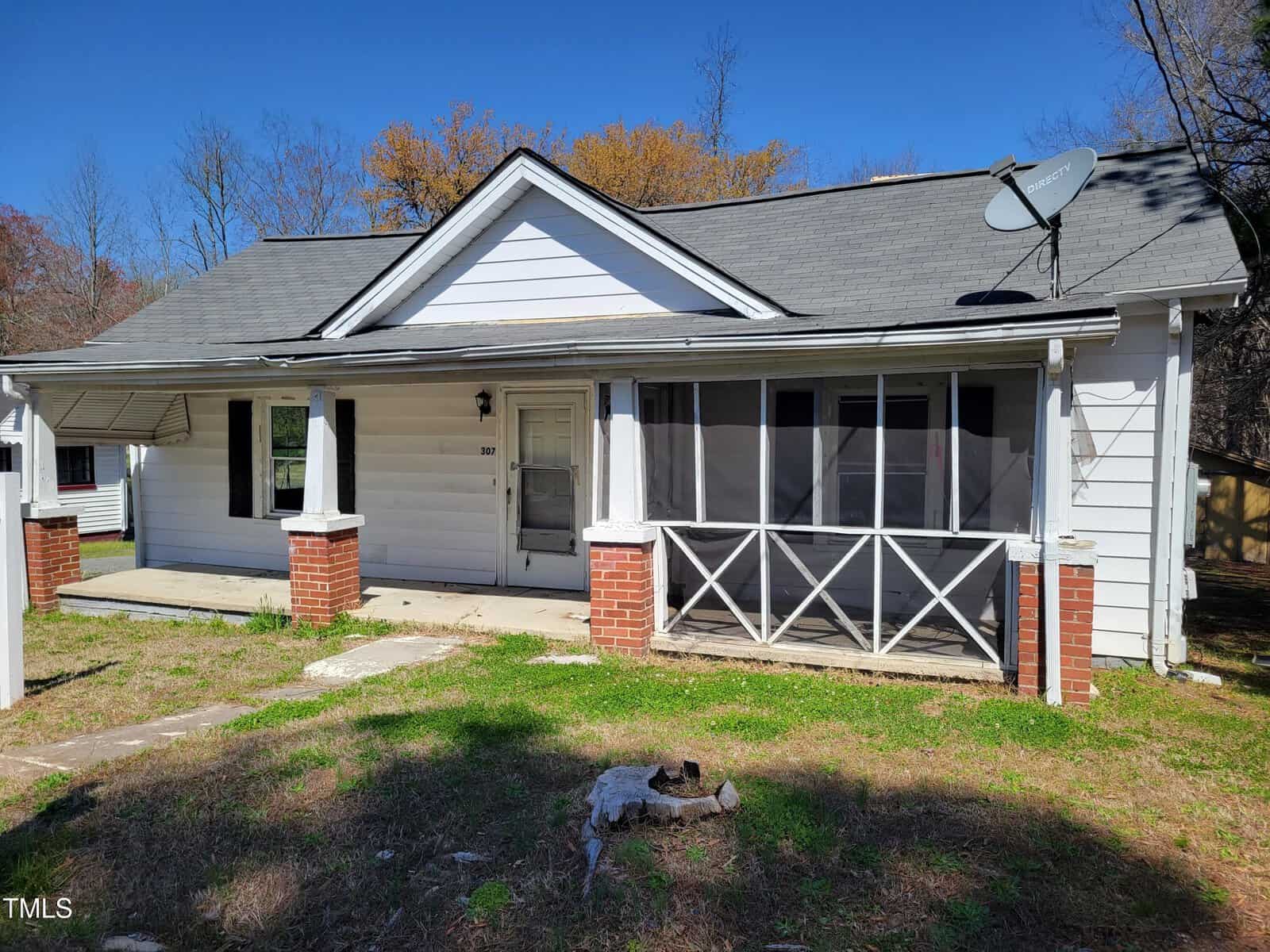 A small, white single-story house with a gray roof and attached porch. The porch has a screen section and brick pillars. A satellite dish is mounted on the roof. The yard is grassy with a few bare spots, and trees are visible in the background.
