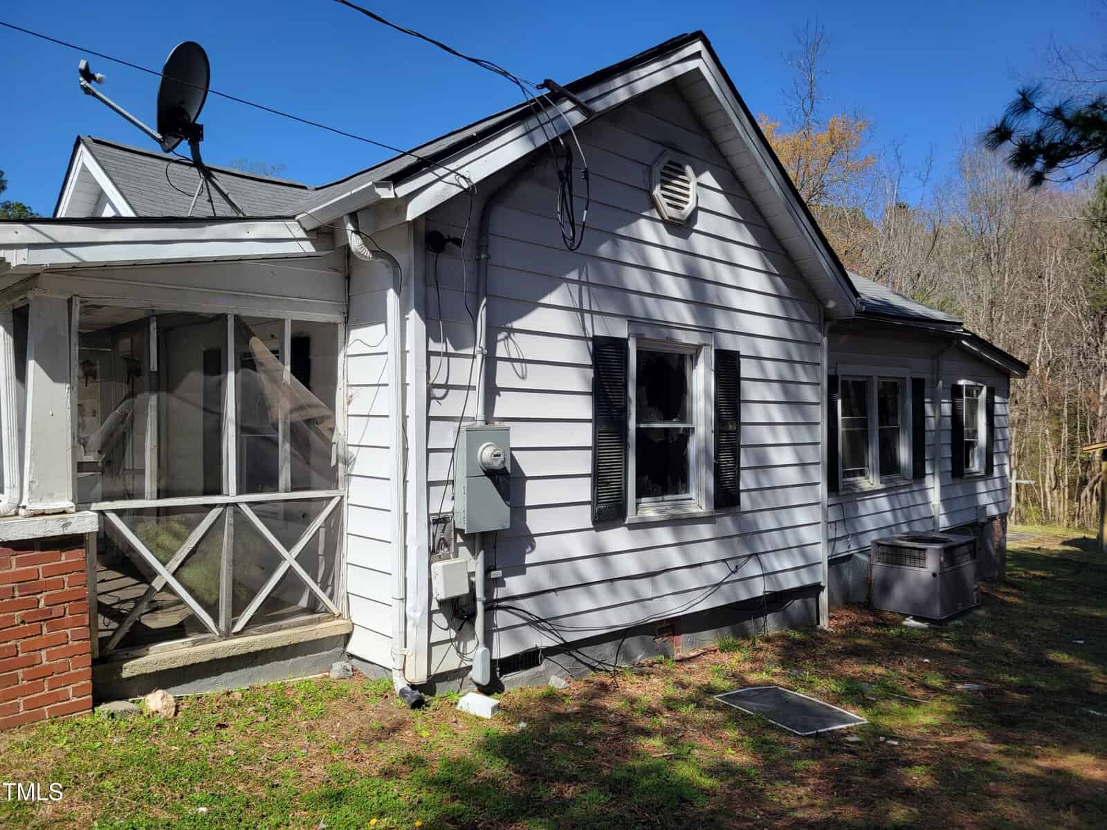 A small, single-story house with white wooden siding, black shutters, and a satellite dish on the roof. There's a screened porch on the left and a wooded area in the background. The yard features some grass and a few trees.