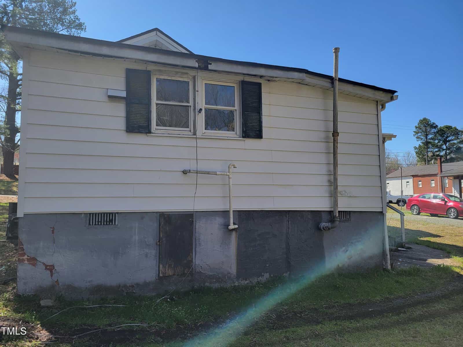 Side view of a small, white house with peeling paint and two windows with black shutters. Pipes and a small metal rail are visible. The foundation is concrete. Grass and another building are in the background.