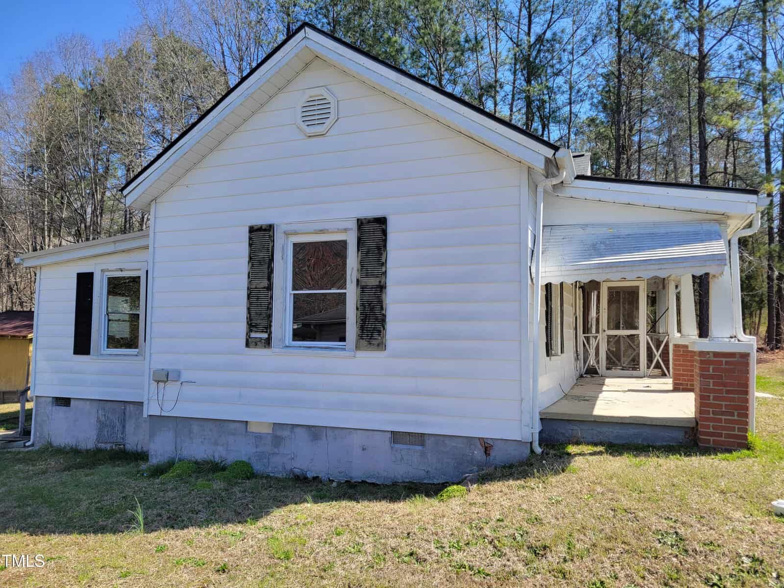 A white single-story house with black shutters and a chimney is shown. There is an attached porch with a white awning. The house is surrounded by grass and trees, under a clear blue sky.