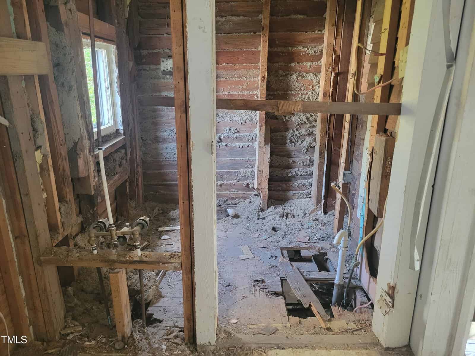 Partially demolished interior of a room, showing wooden studs and insulation in the walls. Exposed plumbing fixtures are visible, and debris is scattered on the floor. A window on the left illuminates the area.