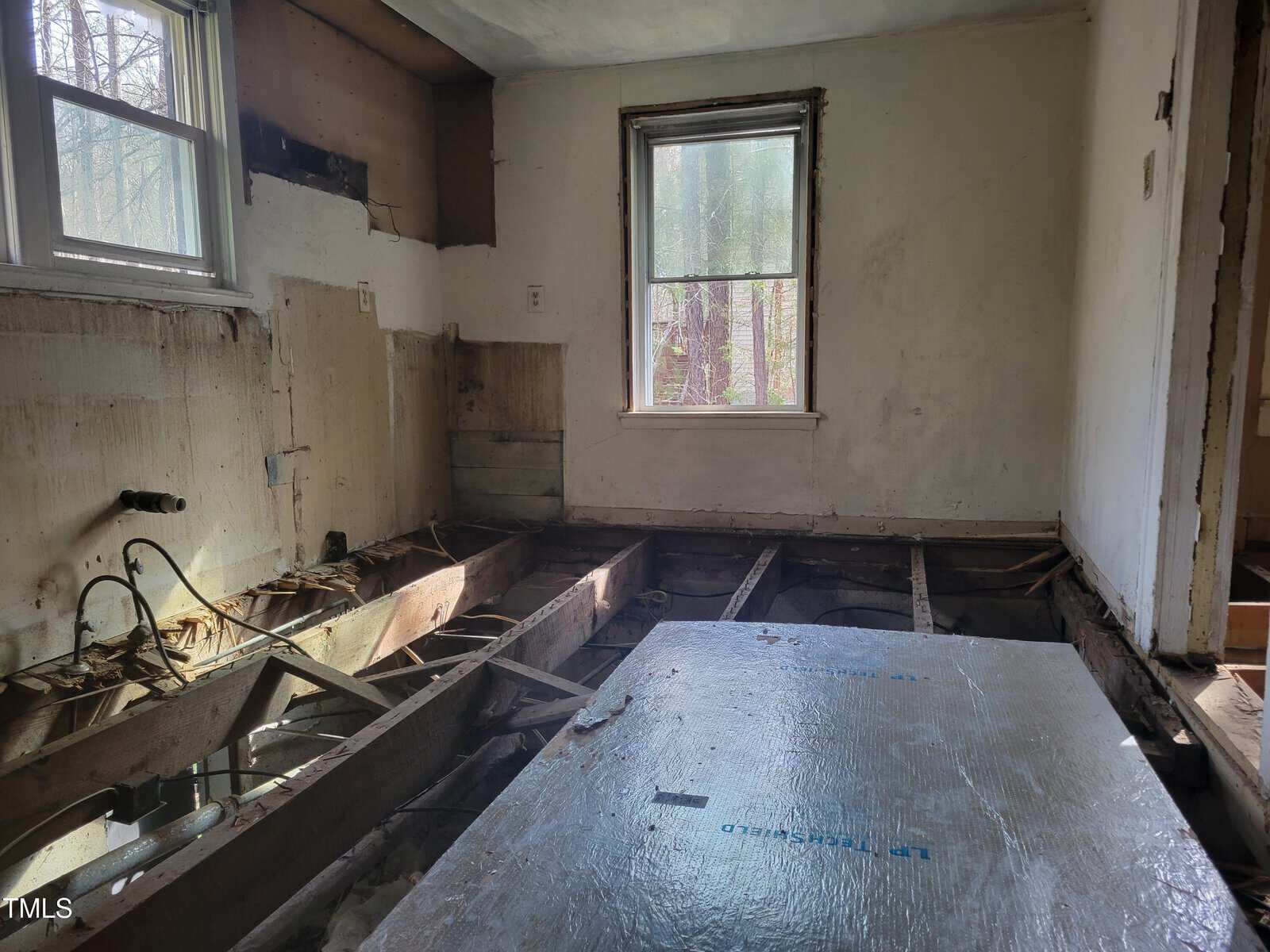 A room under renovation, with exposed floor joists and missing flooring. There is a window with natural light coming in, and another window on the adjacent wall. A large piece of construction material covers part of the floor.
