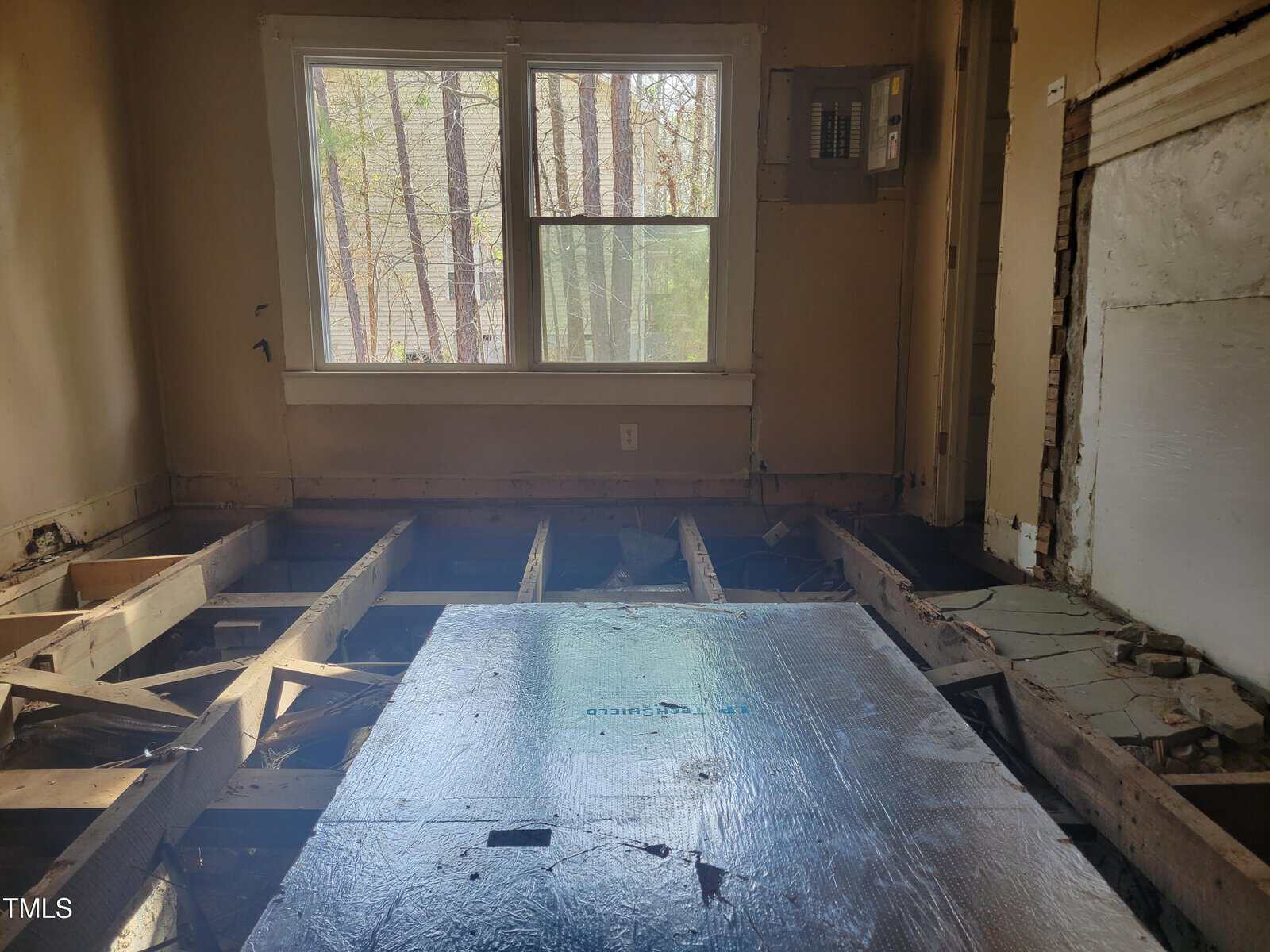 A room under renovation with exposed floor beams and a partially covered plywood sheet. An electrical panel is mounted on the wall. A window provides natural light, showing a view of trees outside. Walls and ceiling are unfinished with visible repairs.