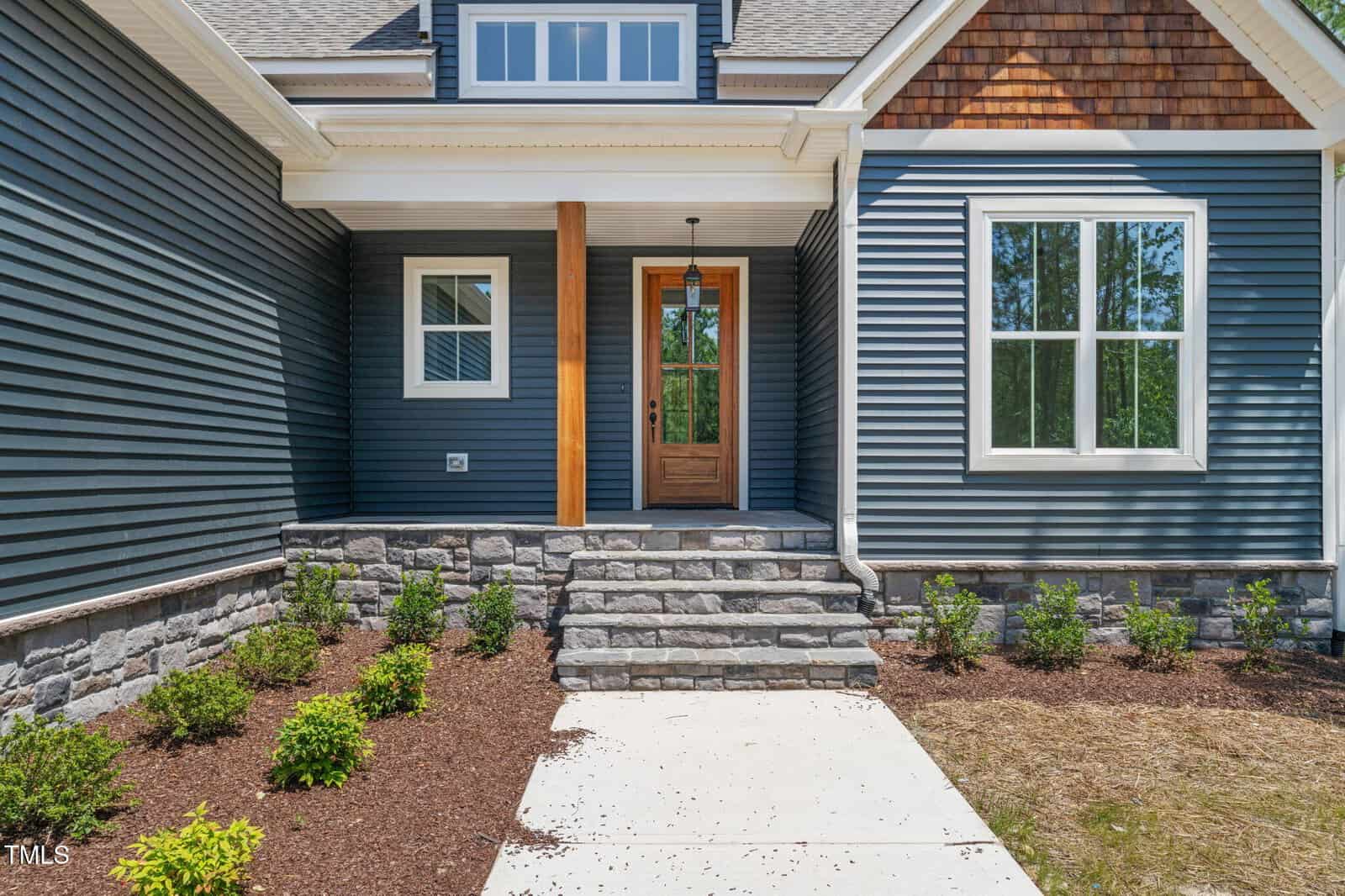A house entrance with gray siding, stone steps leading to a wooden front door with glass panels, a covered porch, and landscaped shrubs along a concrete walkway.