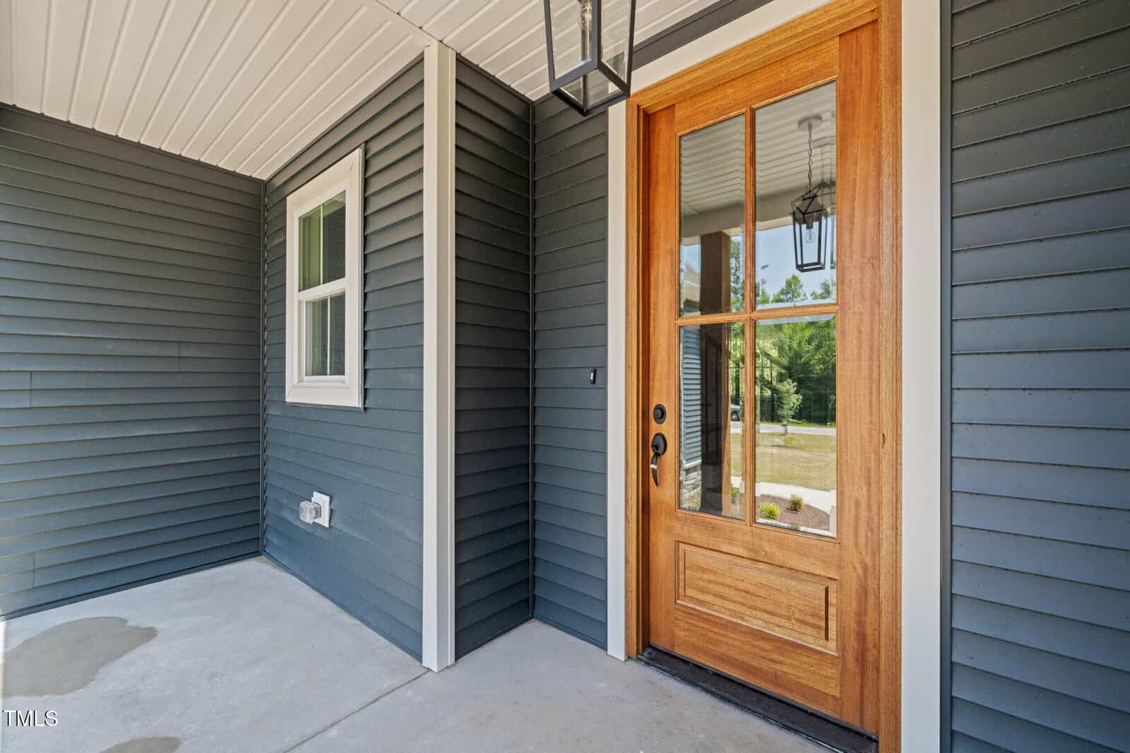 The image shows a covered front porch with blue-gray siding, a single white-framed window, and a wooden front door with glass panels. The porch floor is concrete and a black lantern-style light hangs from the ceiling.