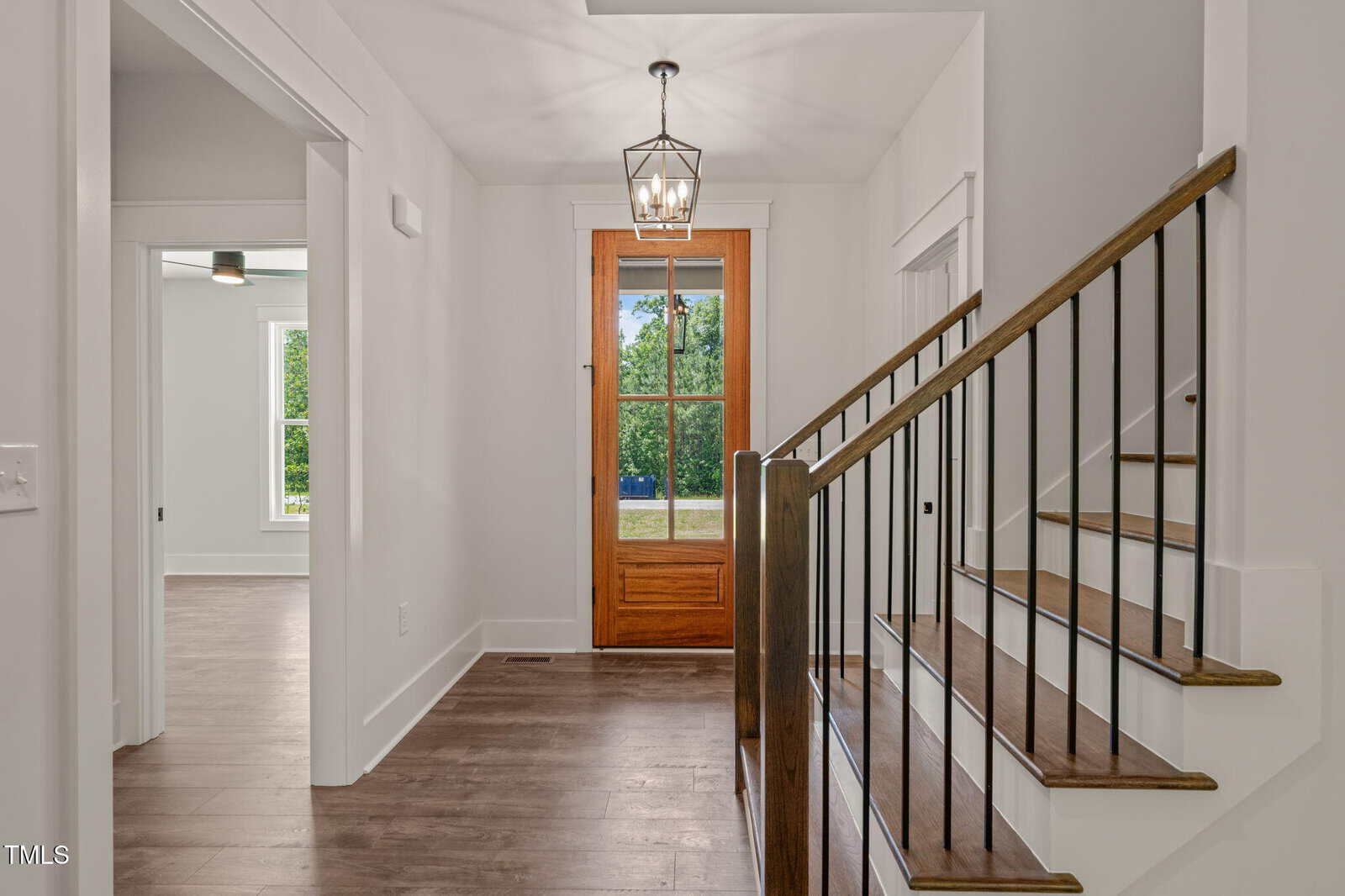 A modern entryway with wood flooring, a wooden front door with glass panels, a metal and wood staircase, white walls, and a geometric ceiling light fixture. A room with large windows is visible to the left.