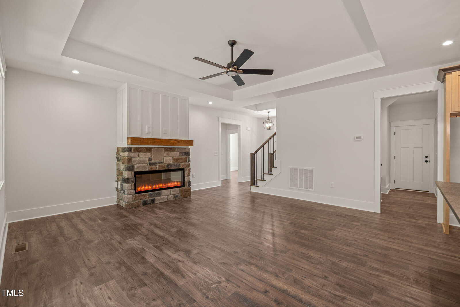 A spacious living room with dark wood floors, a stone fireplace with an electric insert, white walls, a tray ceiling with a ceiling fan, and an open doorway leading to a staircase and entryway.