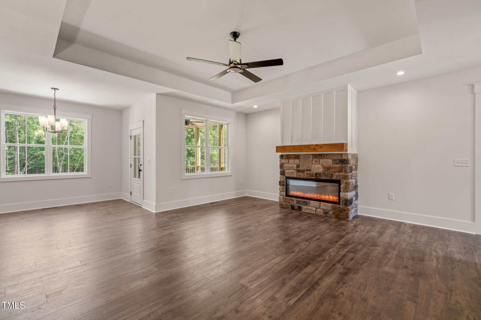 Spacious living area with tray ceiling, ceiling fan, hardwood floors, a stone fireplace with built-in shelf, large windows, and adjoining dining area with chandelier, all in neutral tones.