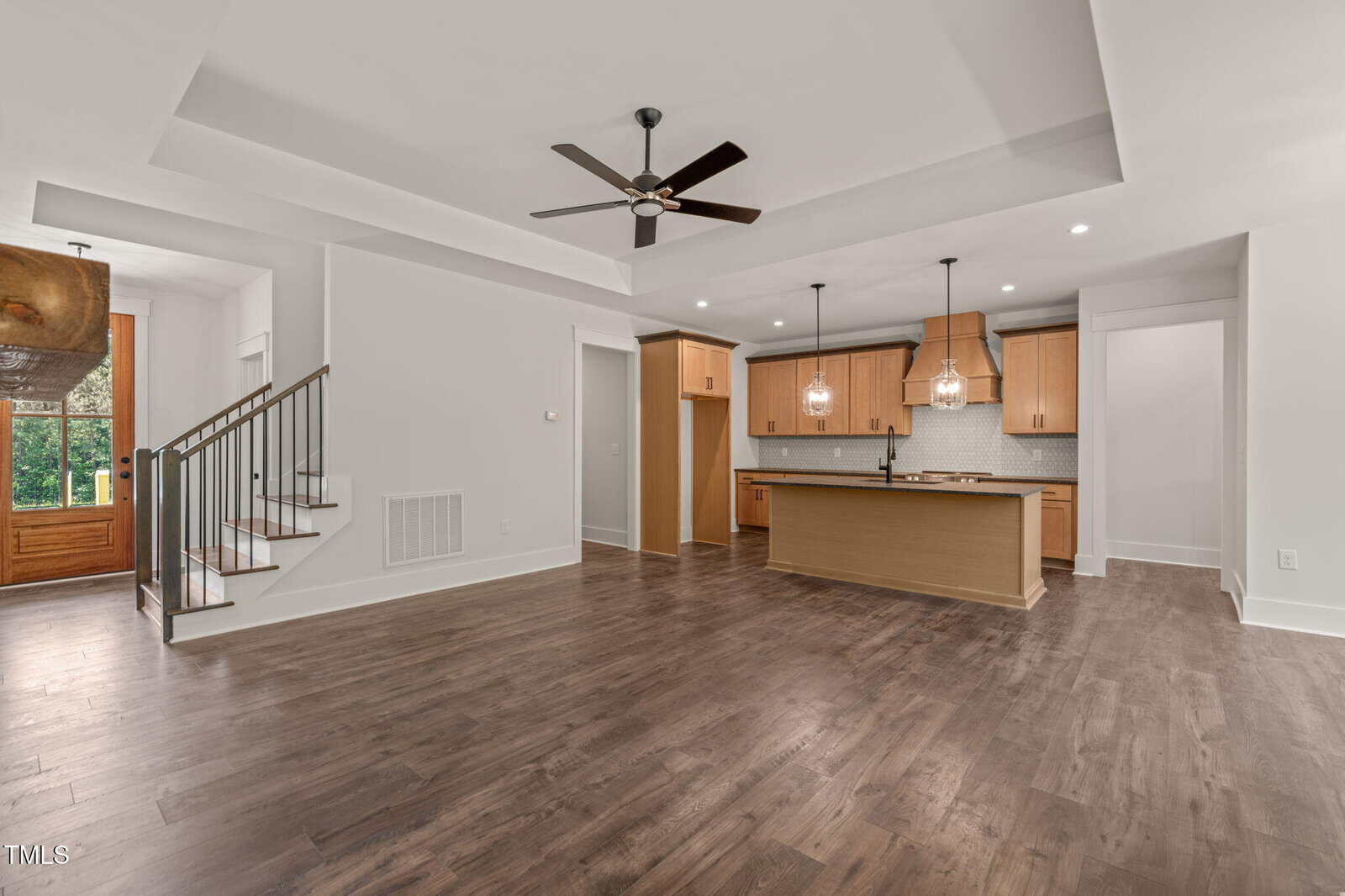 Open-concept room with dark wood floors, light walls, ceiling fan, staircase to the left, and a kitchen area with an island, wooden cabinets, and pendant lights. Natural light comes through a door and window.