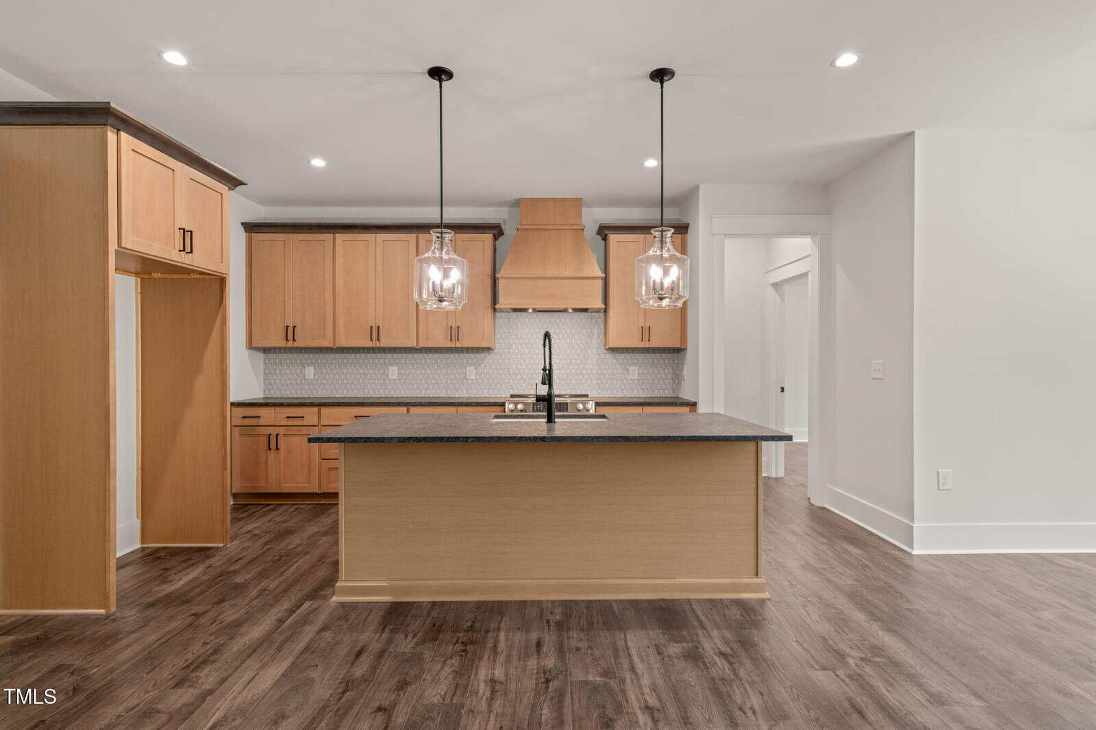 Modern kitchen with wood cabinets, dark countertops, a large island with built-in sink, two pendant lights above, white walls, and dark wood flooring. Stainless steel appliances and a subway tile backsplash are visible.