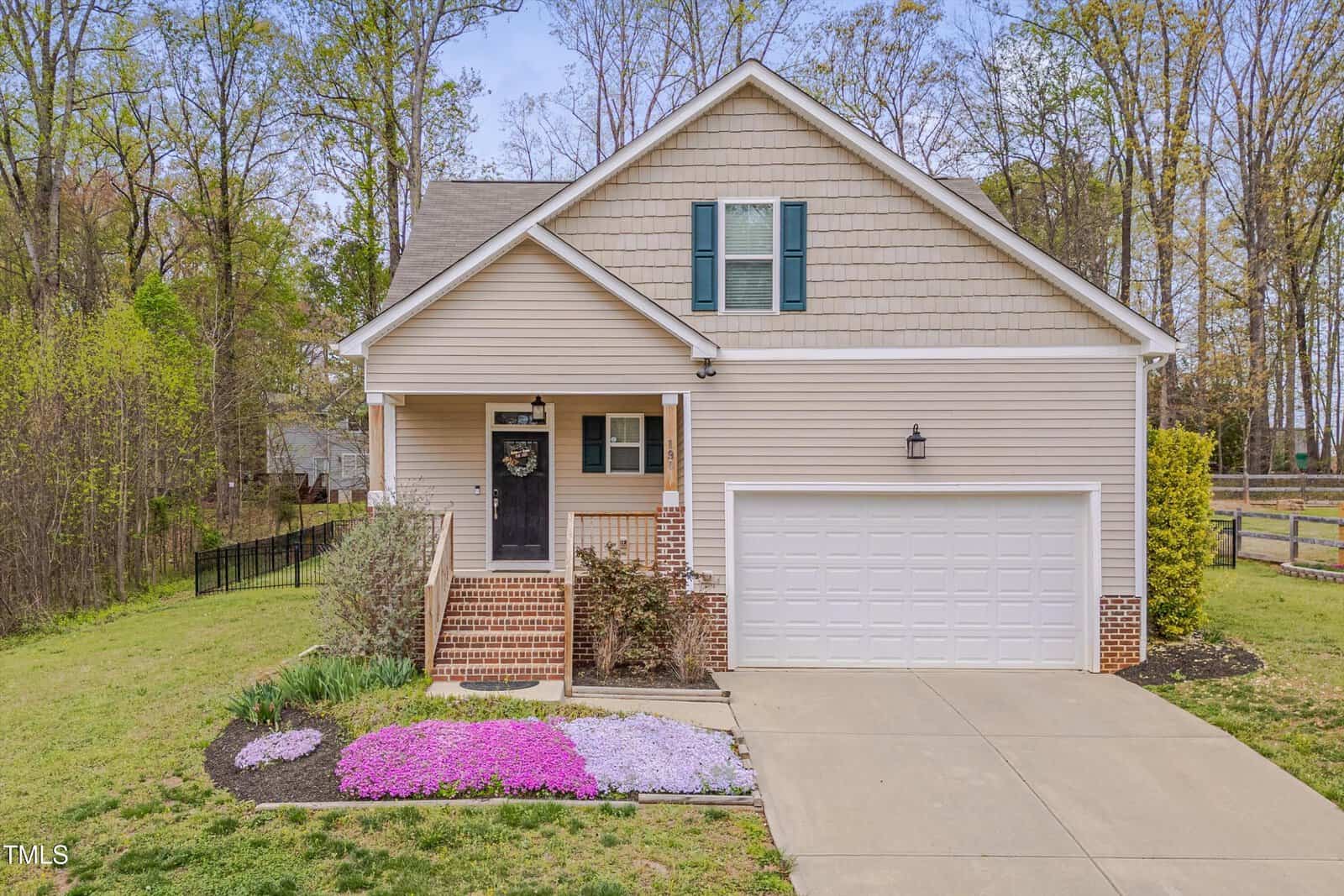 A beige single-family house with green shutters, an attached two-car garage, and a covered front porch. Purple and white flowers border the walkway, and there are trees and grass in the yard.