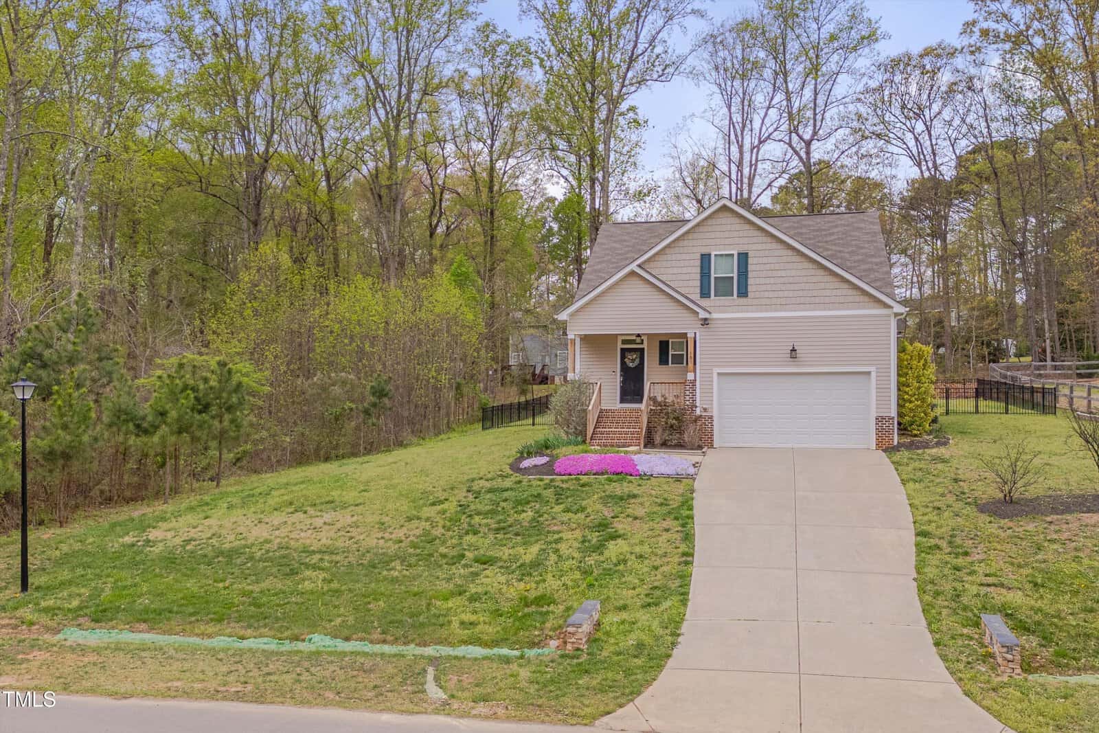 A two-story beige house with a front porch, attached two-car garage, and a long driveway. The yard is partly landscaped with flowers, and the property is bordered by trees and greenery.