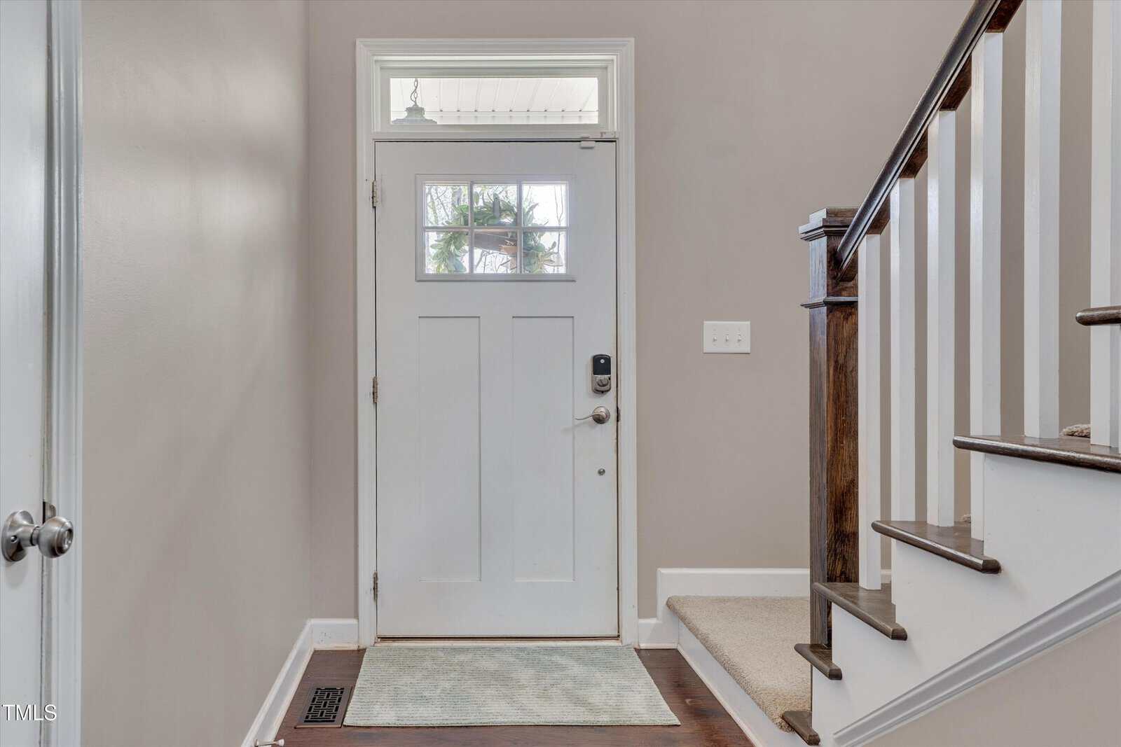 A white front door with four glass panes is next to a staircase with white spindles and a dark wood handrail. The floor is dark wood, and the walls are painted beige. There is a light switch on the wall.