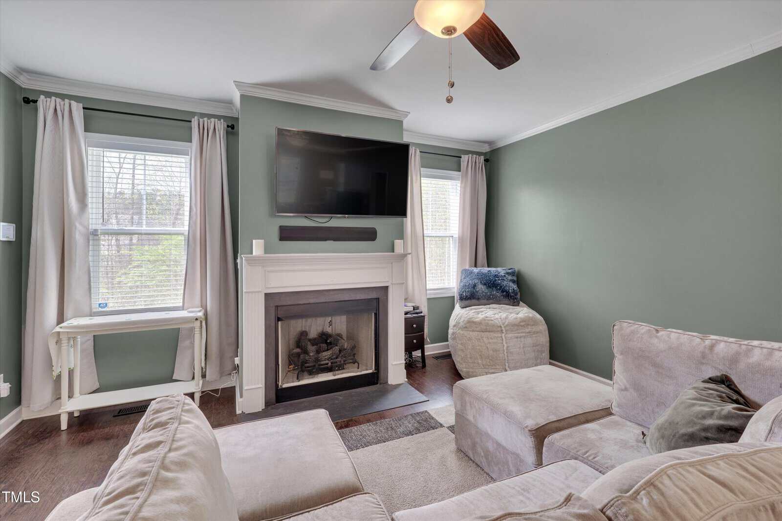A living room with sage green walls, a fireplace with a TV mounted above it, two windows with light curtains, a sectional sofa, a pouf, a side table, and a small white desk against the wall.