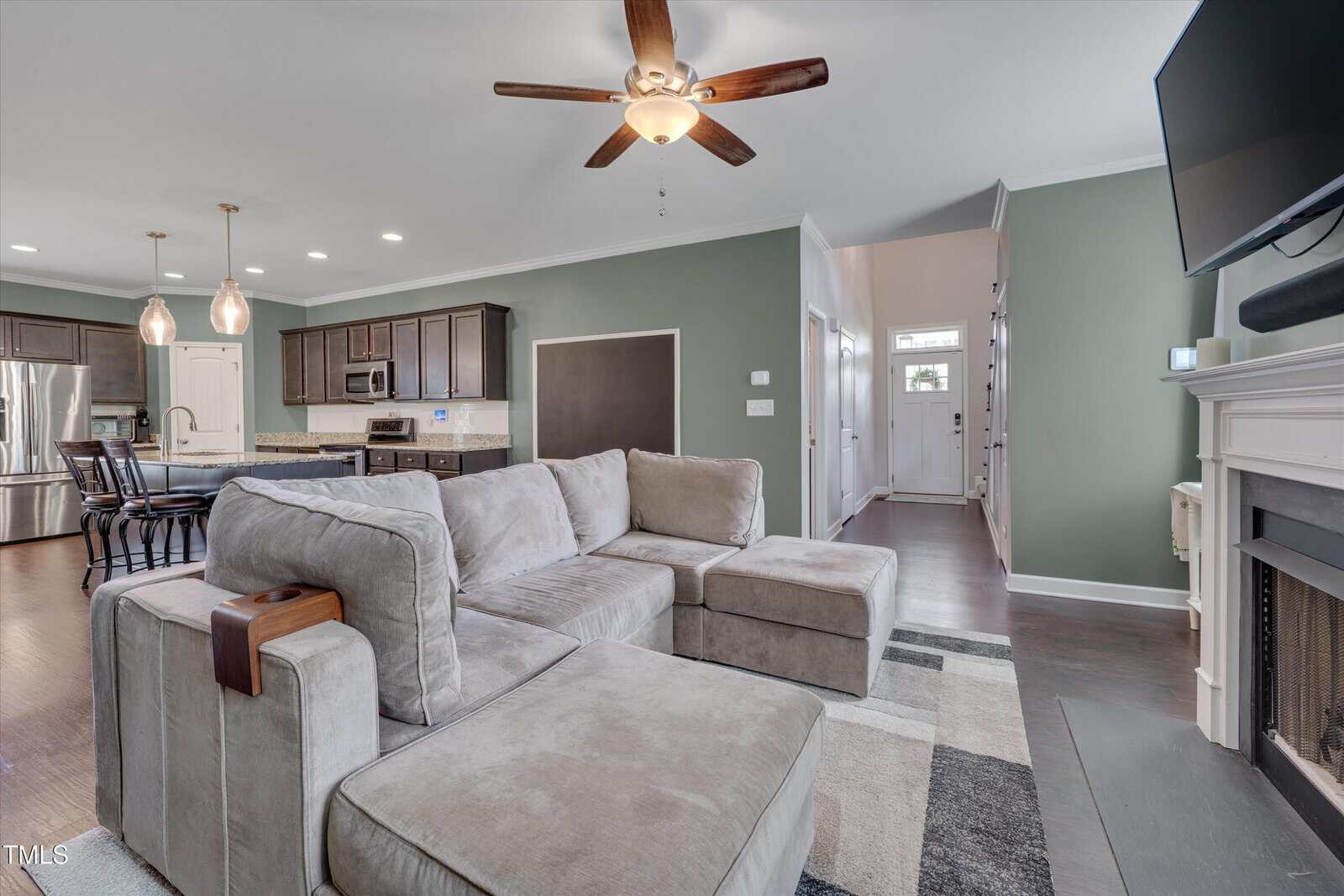Modern open-concept living room and kitchen with gray sectional sofa, dark wood cabinets, stainless steel appliances, ceiling fan, wall-mounted TV, and a fireplace. Hallway and entryway visible in the background.
