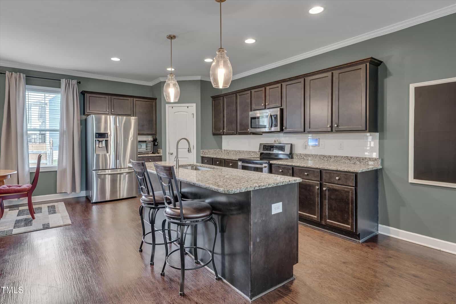 Modern kitchen with dark wood cabinets, stainless steel appliances, granite countertops, an island with two chairs, pendant lights, and hardwood floors. A dining table is partially visible near a window with blinds and curtains.