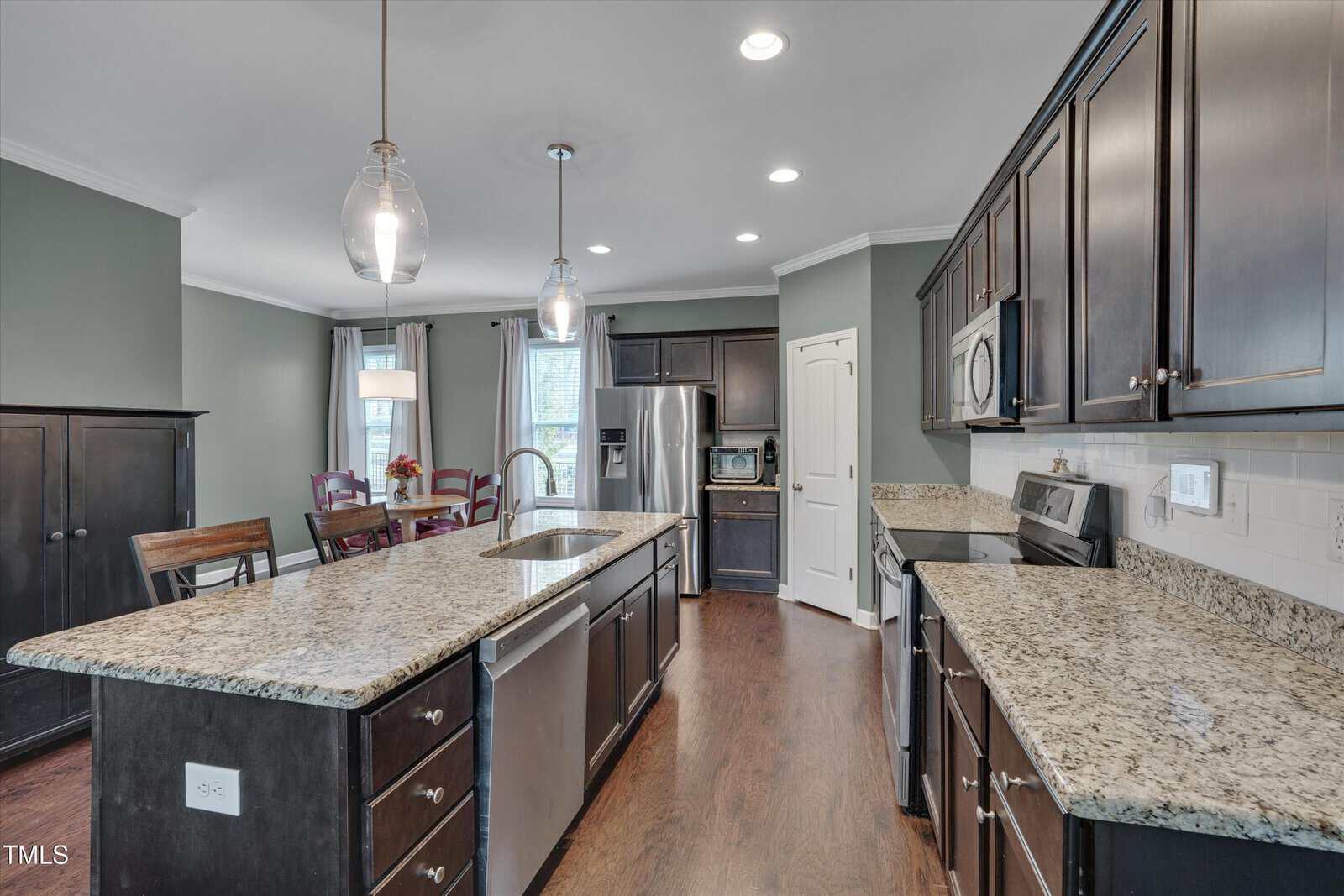 Modern kitchen with dark wood cabinets, granite countertops, stainless steel appliances, an island with a sink and dishwasher, pendant lighting, and adjacent dining area with a table and chairs. Hardwood flooring throughout.