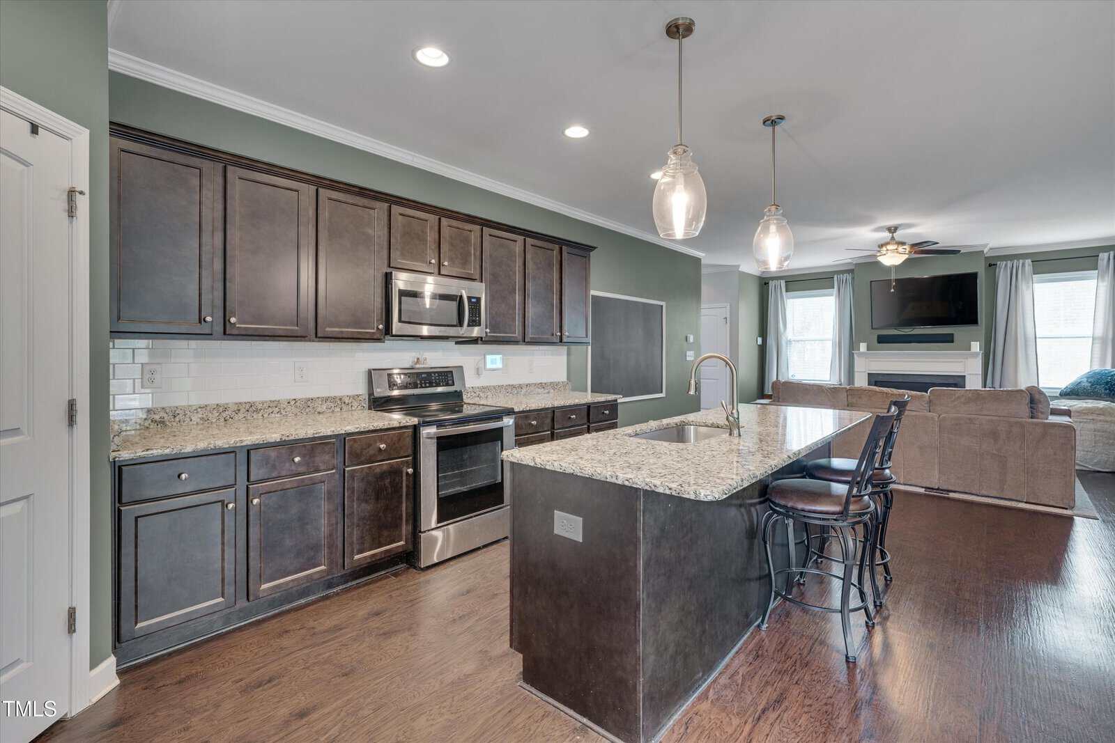 Modern kitchen with dark cabinets, stainless steel appliances, granite countertops, island with sink and barstools, pendant lights, and open layout leading to a living room with a wall-mounted TV and large windows.