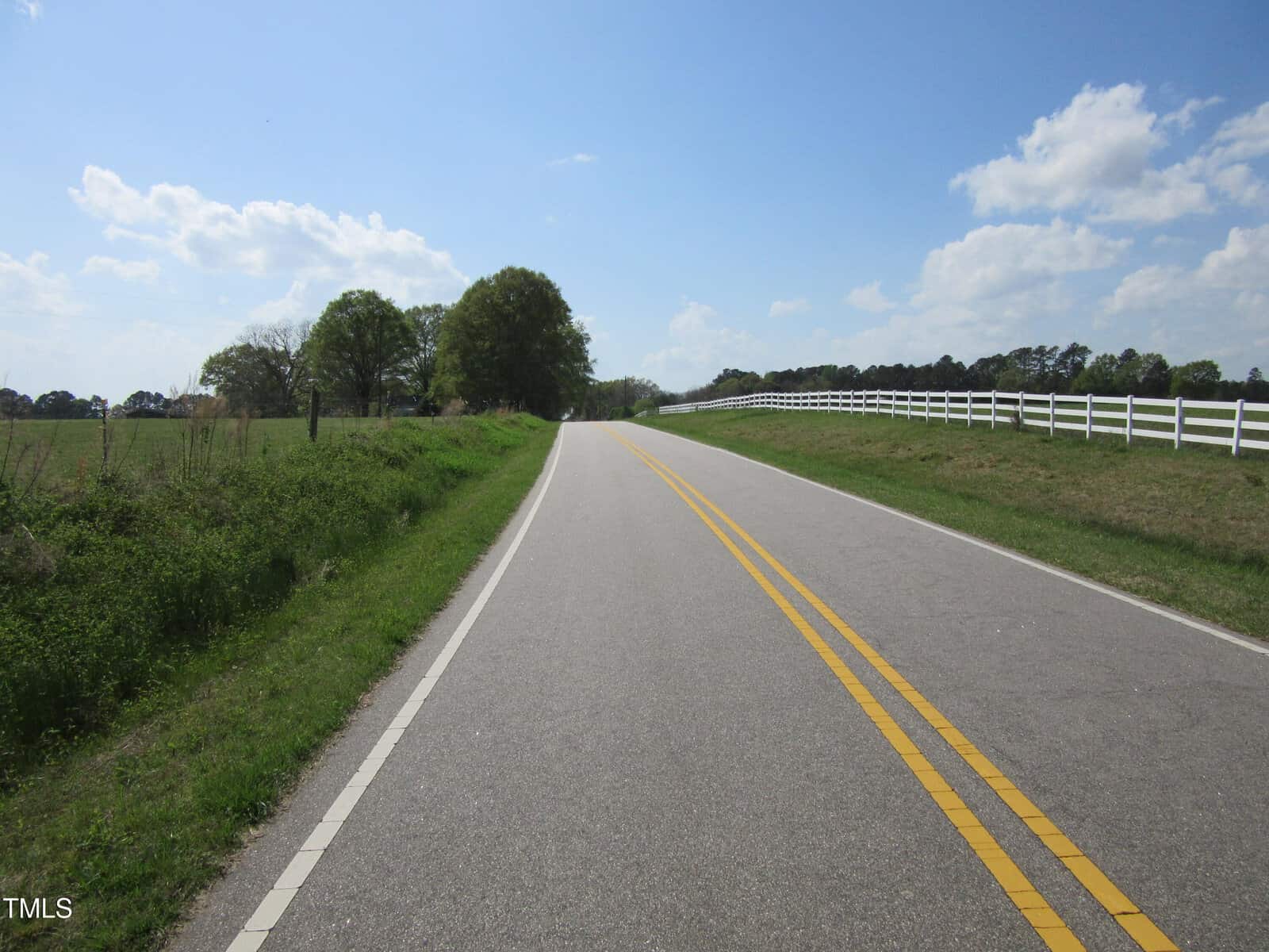 A two-lane paved rural road with double yellow lines curves gently ahead. Grass, a tree line, and a white fence border the road under a partly cloudy blue sky.
