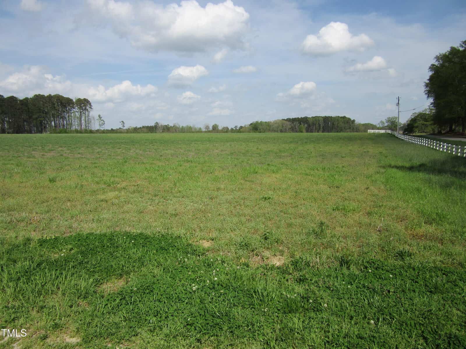 A wide, open grassy field extends toward the horizon with a line of trees on the left and right sides. A white fence runs along a road to the right, marking the edge of Tract 1 Pine Ridge Road in Zebulon. The sky is partly cloudy.