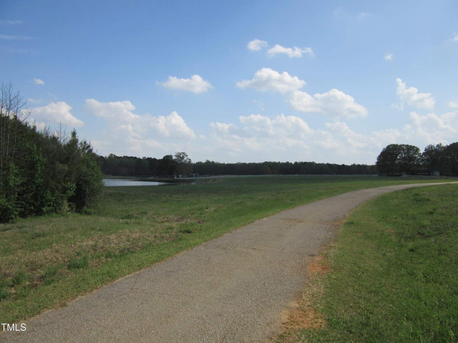 A paved path curves through a grassy field under a blue sky with scattered clouds. Trees line the horizon of Tract 2 Pine Ridge Road, and a small body of water is visible on the left side of the image in Zebulon.