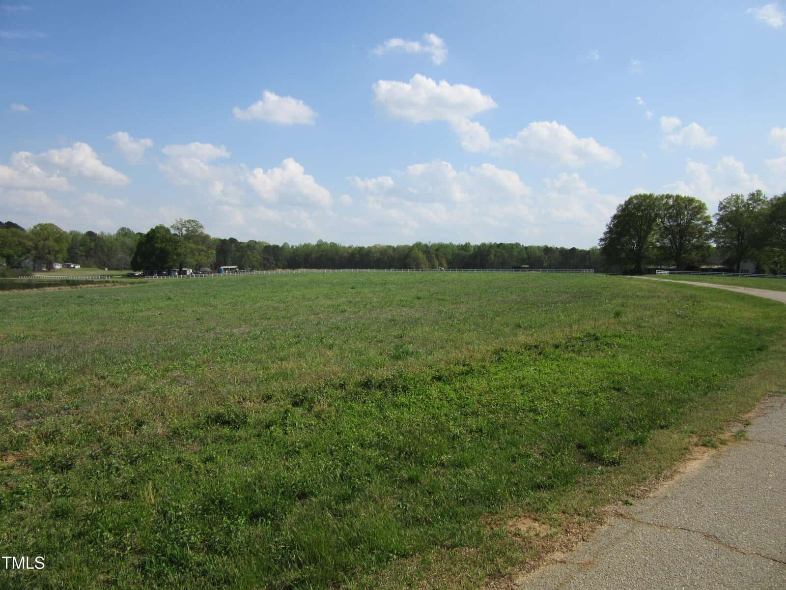 A large, grassy field bordered by trees under a partly cloudy sky. A paved path runs along the right side, and a few buildings are visible in the distance to the left.