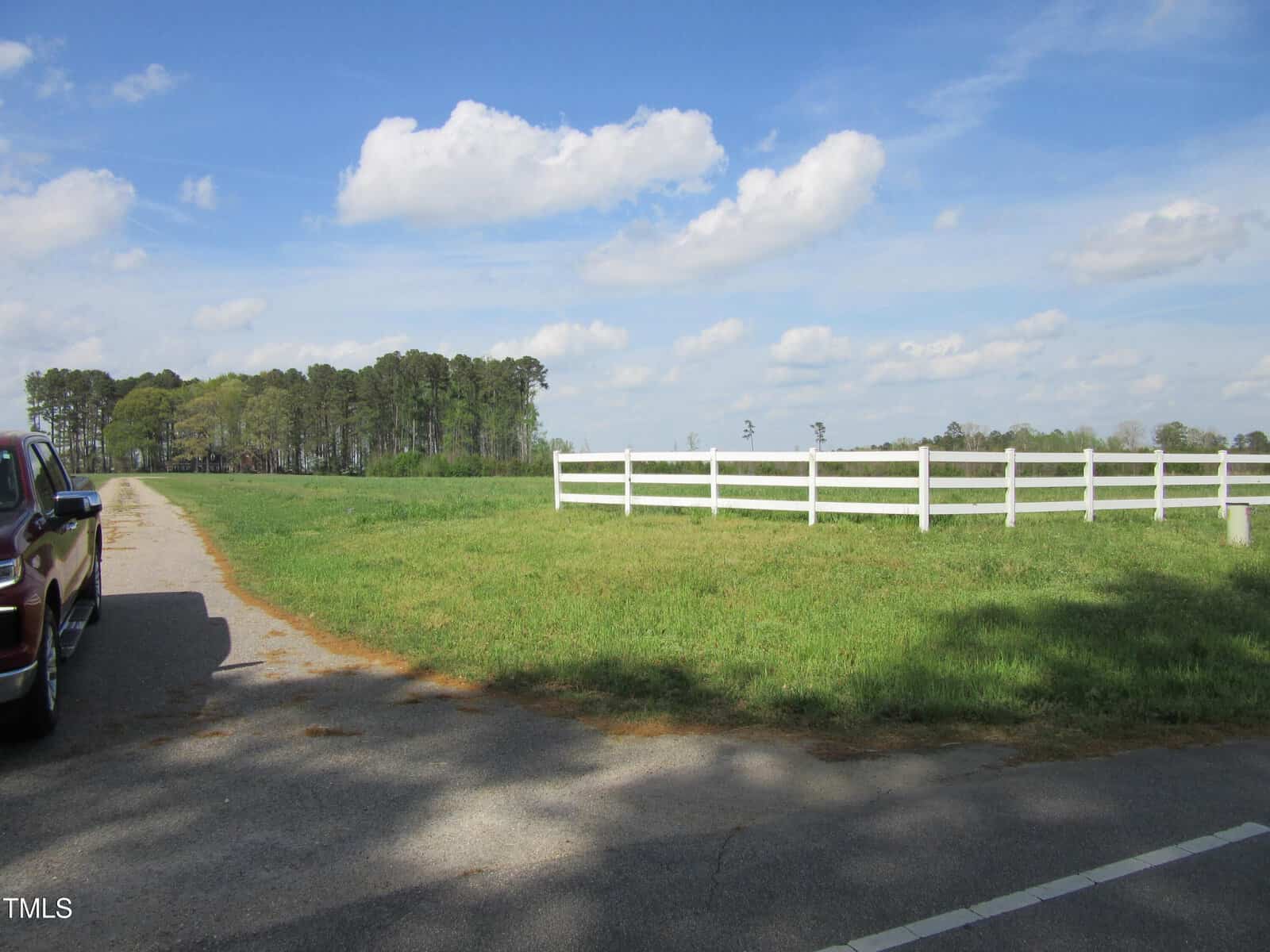 A rural scene with a grassy field, a white wooden fence, a red vehicle partially visible on the left, a paved road, scattered trees in the background, and a blue sky with clouds.