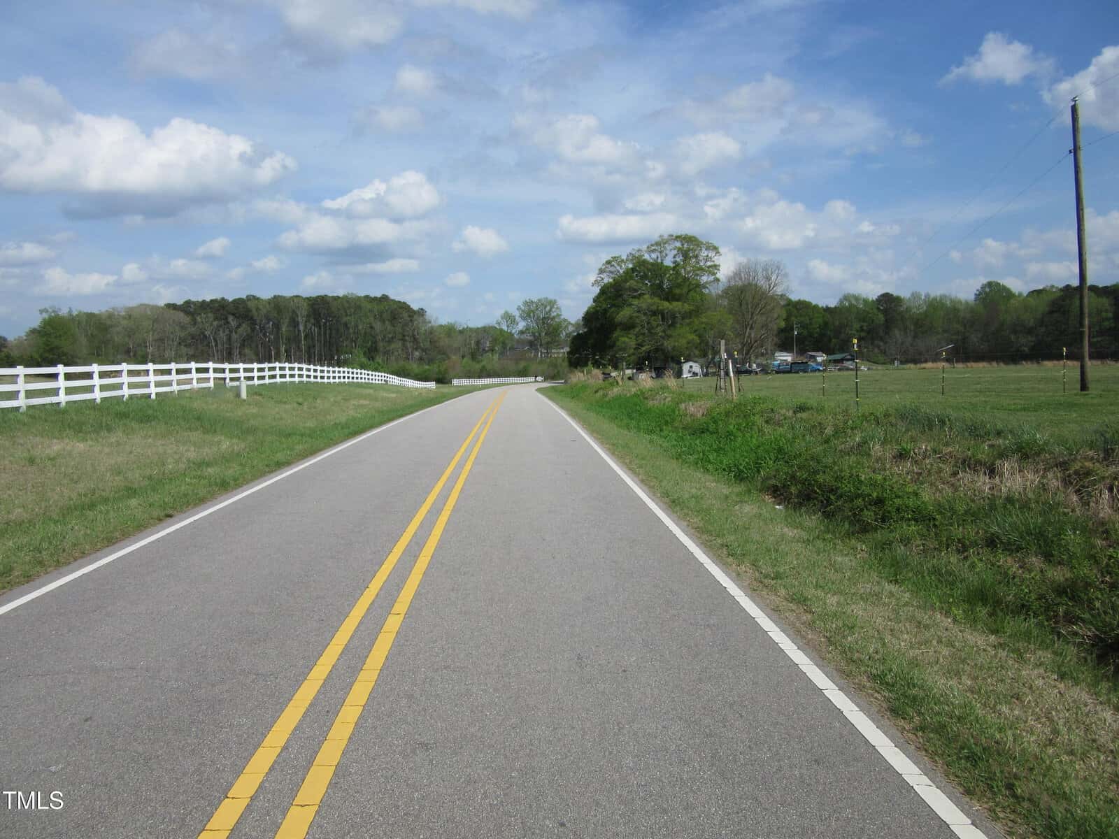 A two-lane country road with double yellow lines runs through a rural area, bordered by a white fence and grassy fields under a partly cloudy sky. Trees and a few buildings are visible in the background.