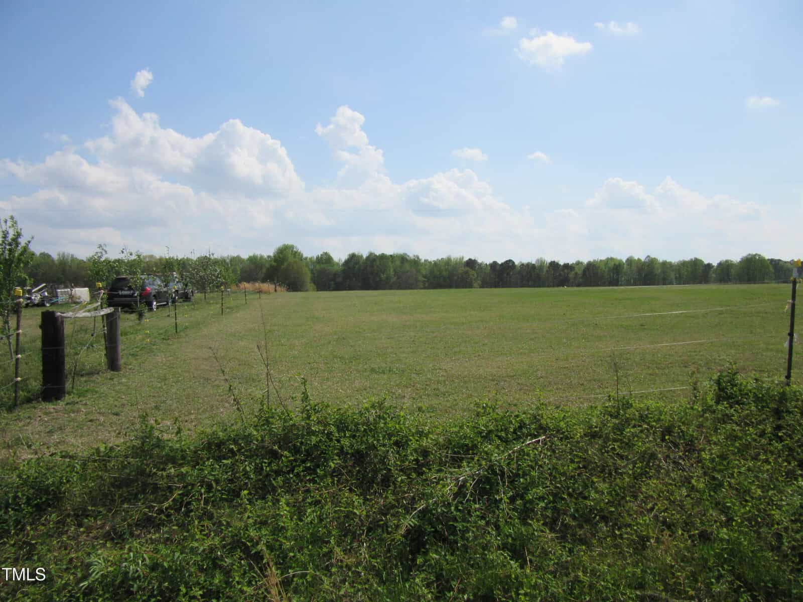 A wide, open grassy field is bordered by a fence and trees in the distance under a partly cloudy sky. A few vehicles are parked to the left near the fence. The image appears to be taken in a rural area along Tract 4 Pine Ridge Road, near Zebulon.