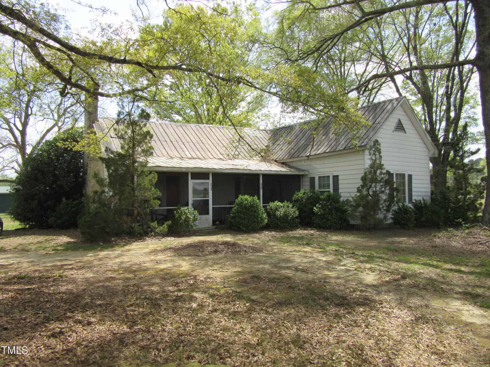 A small, single-story white house with a metal roof and screened porch is nestled among tall trees and bushes on Tract 5 Pine Ridge Road in Zebulon. The yard is mostly bare ground with scattered leaves and shade from the overhanging tree branches.