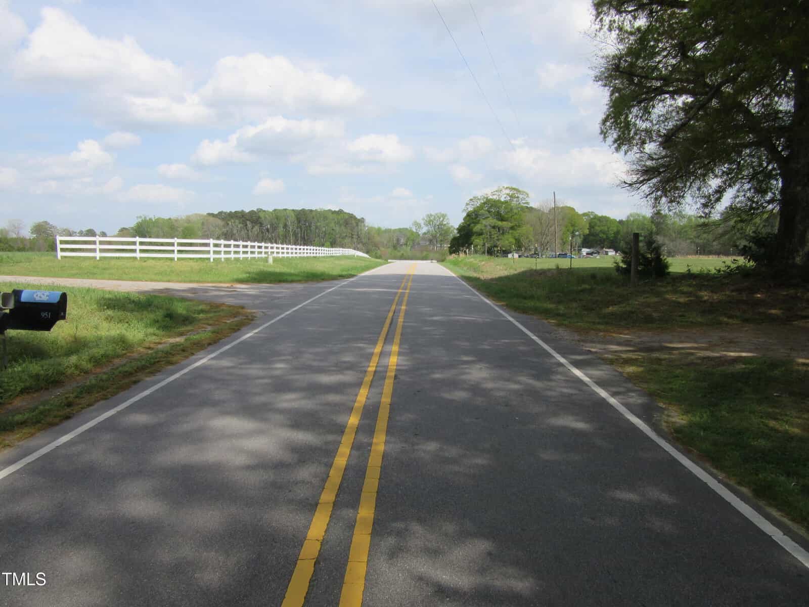 A paved rural road with double yellow lines runs straight ahead, bordered by grass, a white fence on the left, and trees on both sides under a partly cloudy sky. A black mailbox is visible on the left.
