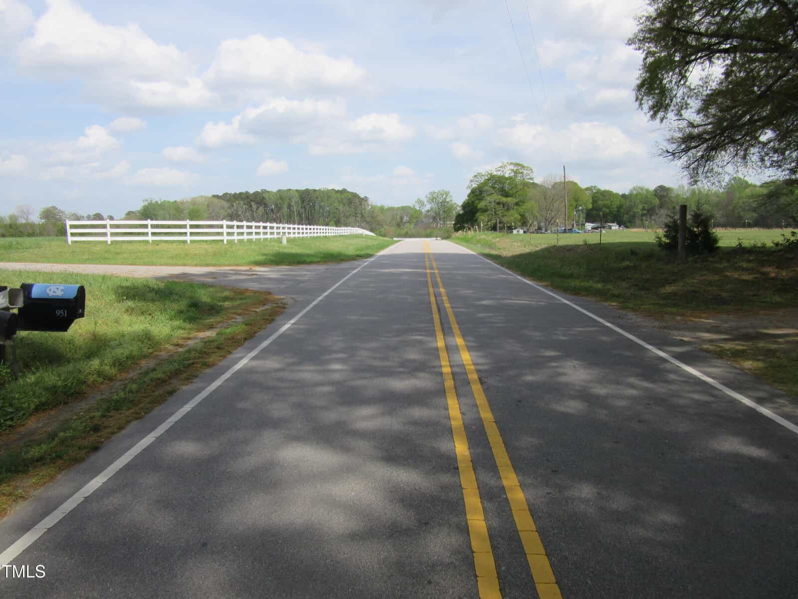 A paved road with double yellow lines runs through a rural area, bordered by grassy fields, a white fence on the left, trees in the distance, and a mailbox on the left foreground under a partly cloudy sky.