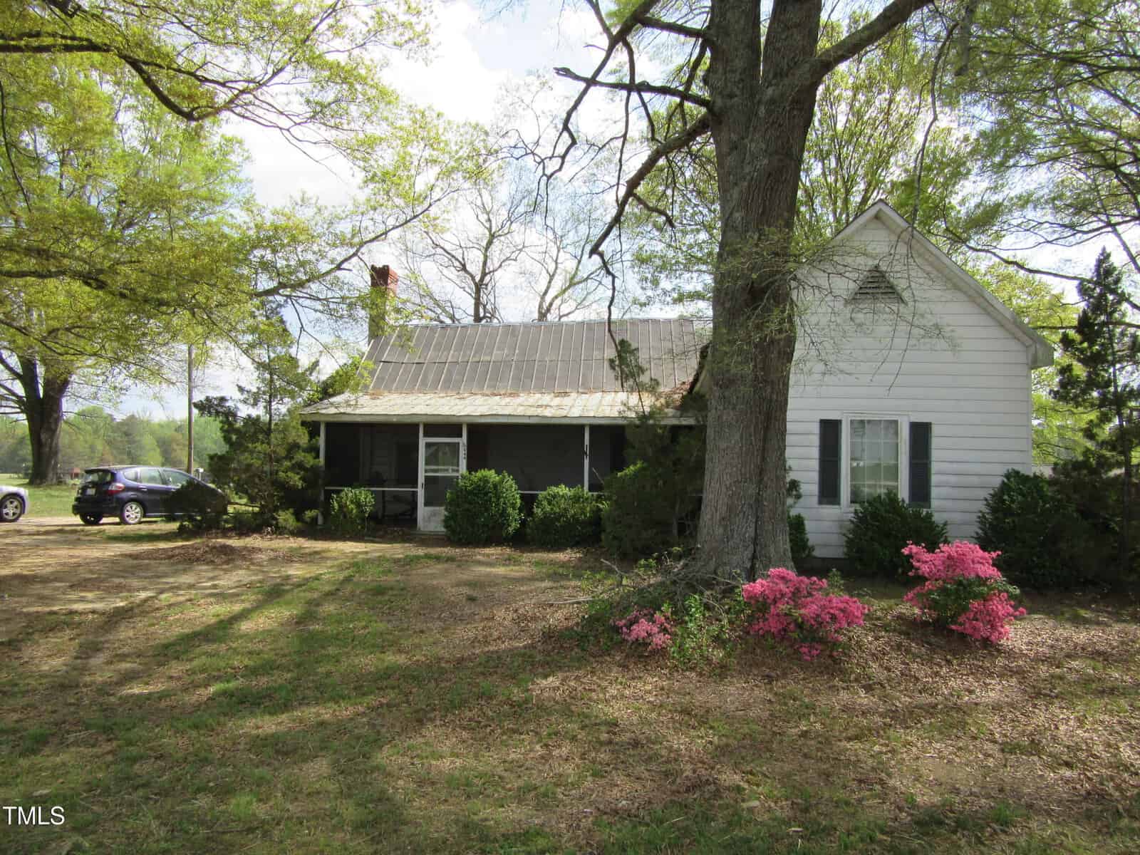A white, single-story house with a metal roof sits behind trees and bushes. Pink flowers bloom in the foreground, and a black car is parked to the left. The yard is grassy with scattered shade from large trees.