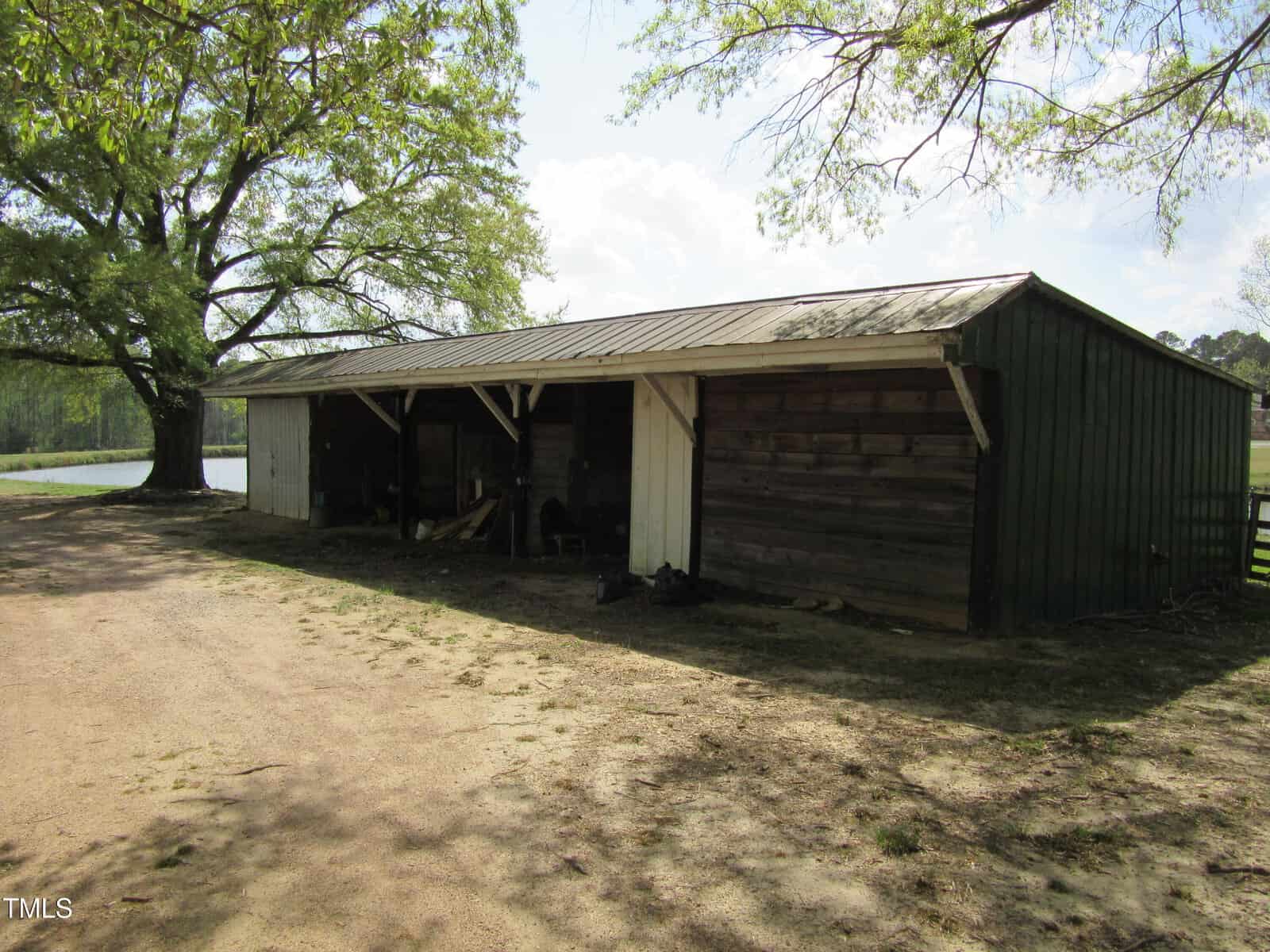 A rustic wooden shed with a metal roof sits beside a dirt path under a large tree, with fields and a pond visible in the background on a sunny day.