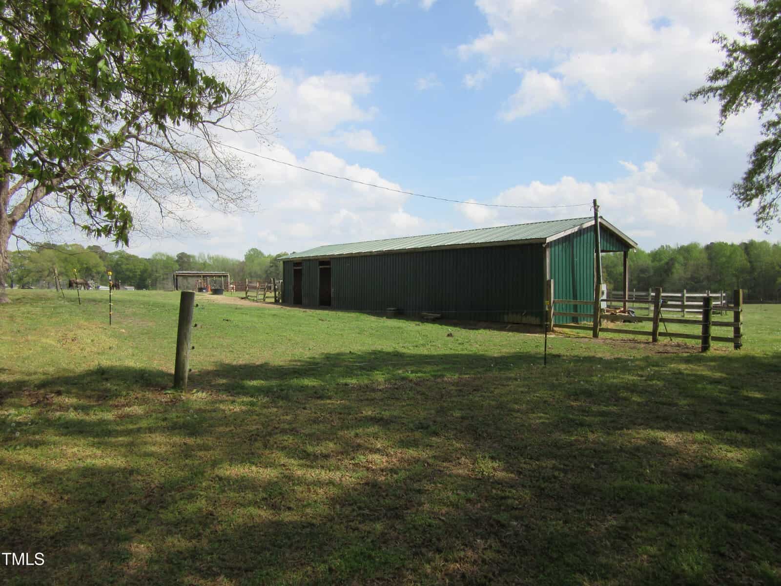 A long green metal barn stands in a grassy field with wooden fences, under a partly cloudy sky. Trees and open pasture are visible in the background.