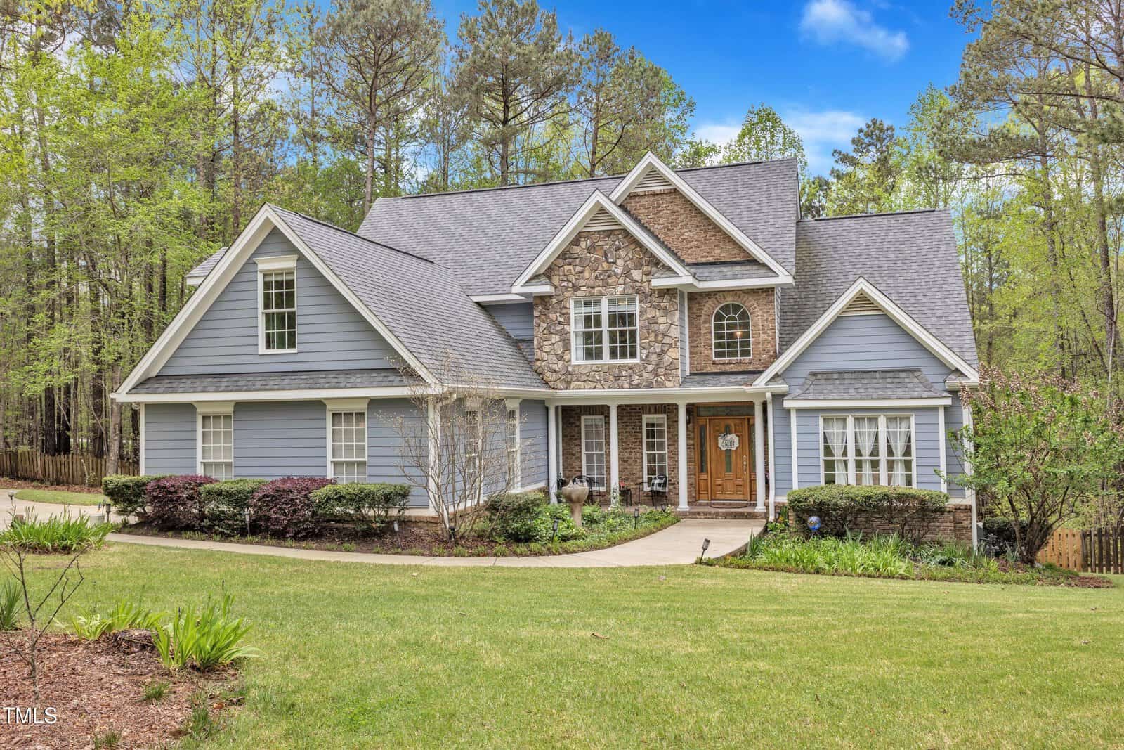 A charming two-story house at 250 Highview Drive in Youngsville boasts blue siding and stone accents, framed by a serene woodland backdrop. It features a gray roof, double front doors adorned with a wreath, and symmetrical windows. A curved walkway through landscaped shrubs leads to the entrance.