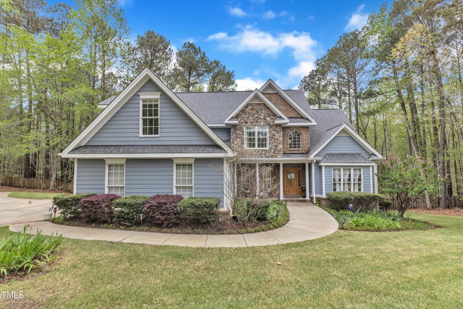 A two-story house with blue siding and stone accents is surrounded by a circular driveway and landscaped with shrubs. The roof is gray, and large windows are present. The property is bordered by trees, and the sky is clear with some clouds.