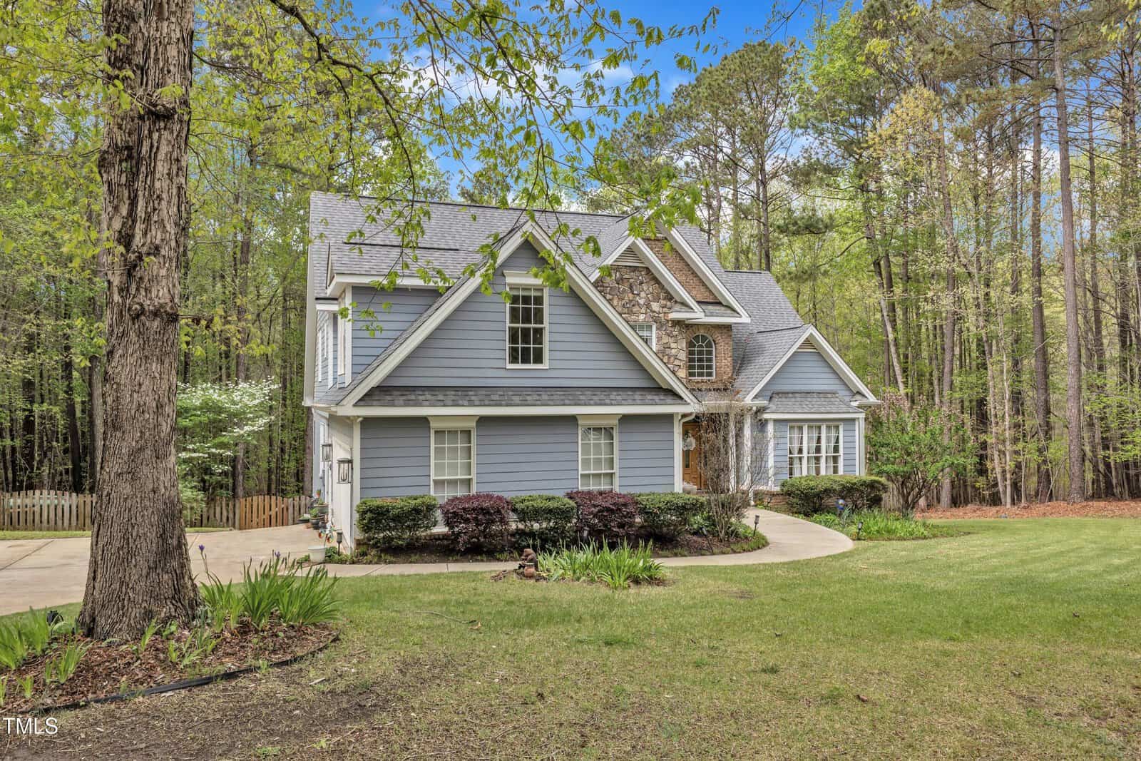 A two-story house with gray siding and stone accents, surrounded by trees. It features a sloped roof and manicured shrubs along the driveway. The yard is spacious with a well-maintained lawn. A wooden fence and pathway are visible to the side.