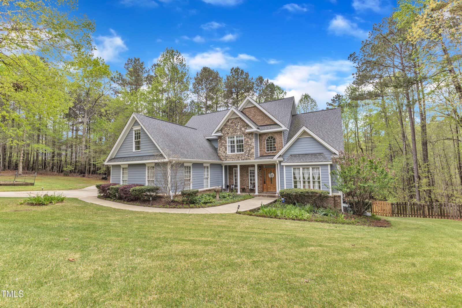 A two-story house with a combination of stone and gray siding exterior, featuring a gabled roof and a wooden front door. The house is surrounded by a lawn, trees, and a paved driveway, set against a backdrop of a forest and a blue sky.