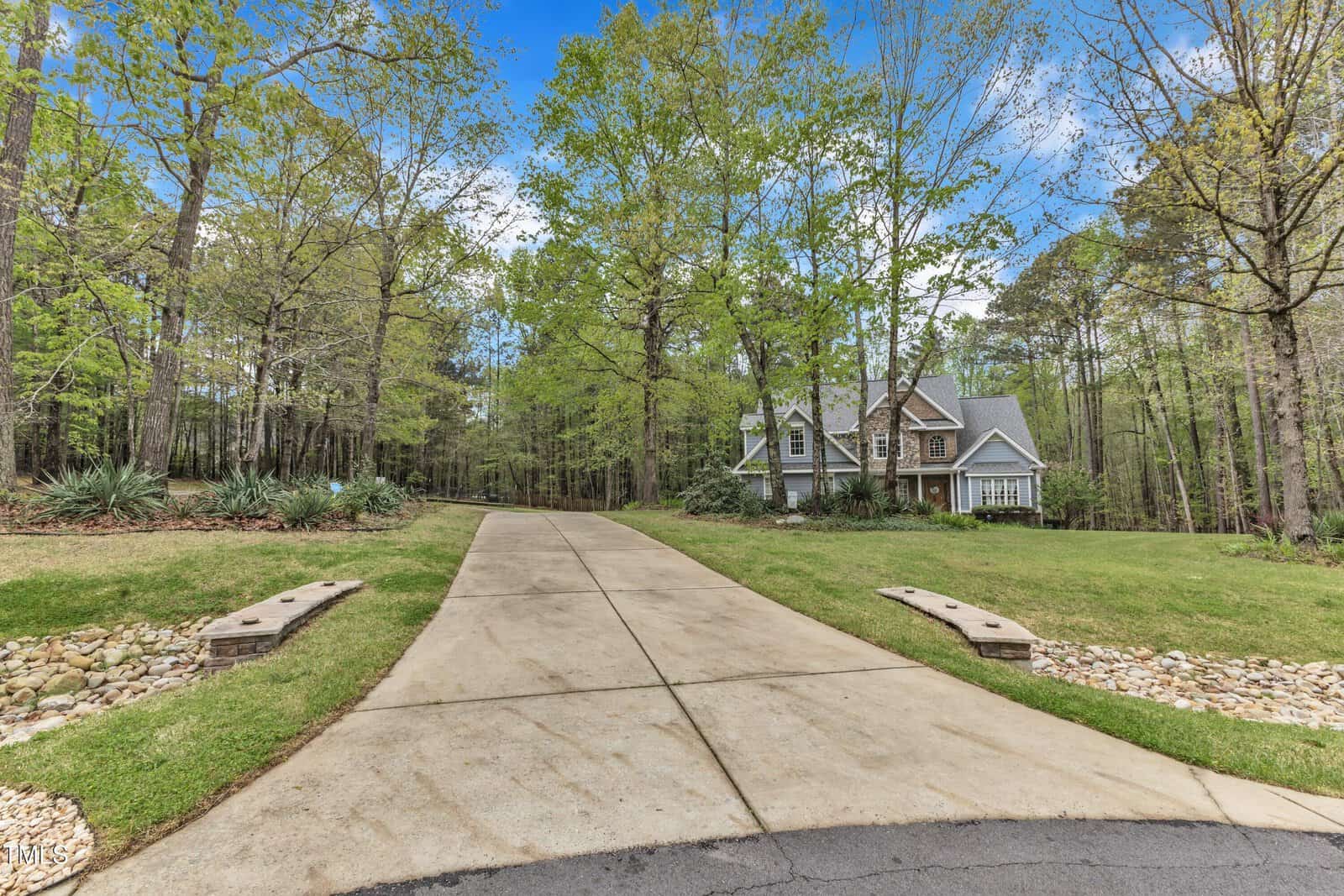 A long driveway leads to a two-story house surrounded by trees. The house has a gray facade with white trim, and a steeply pitched roof. The landscape is grassy with some low shrubbery lining the driveway. The sky is clear and blue.