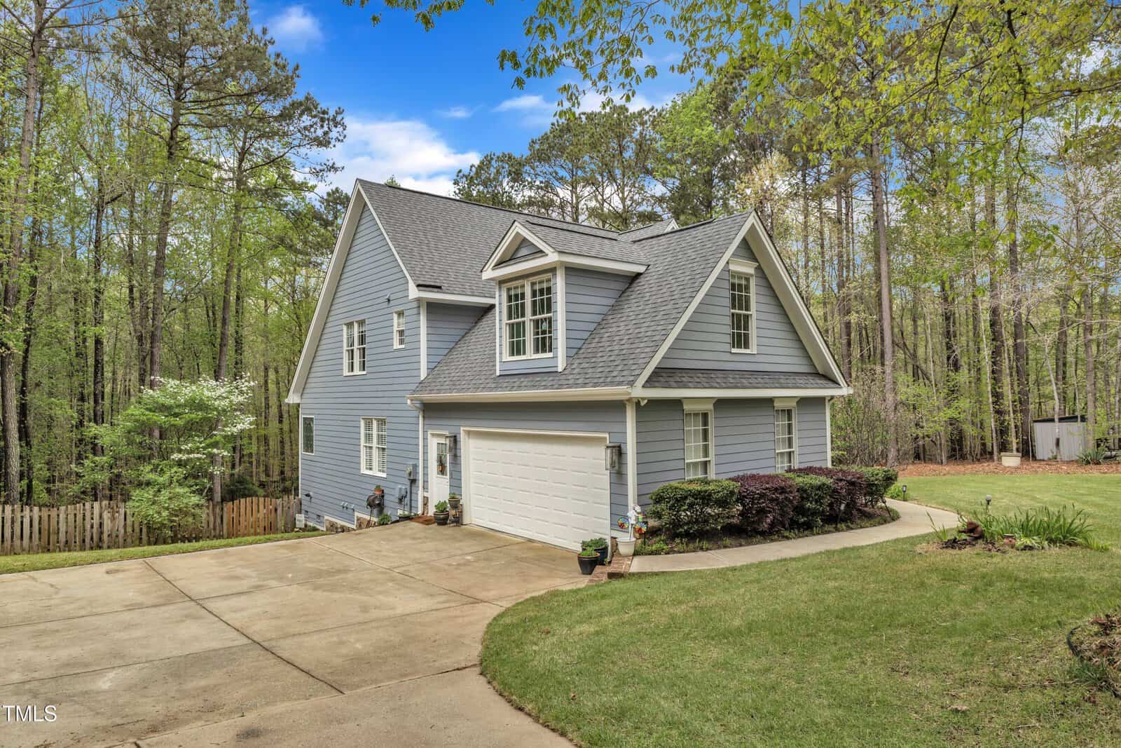 A two-story blue house with a gray roof, surrounded by trees. It features a white garage door and a concrete driveway. A well-maintained lawn and shrubbery are visible in the front yard. A wooden fence runs along the left side.