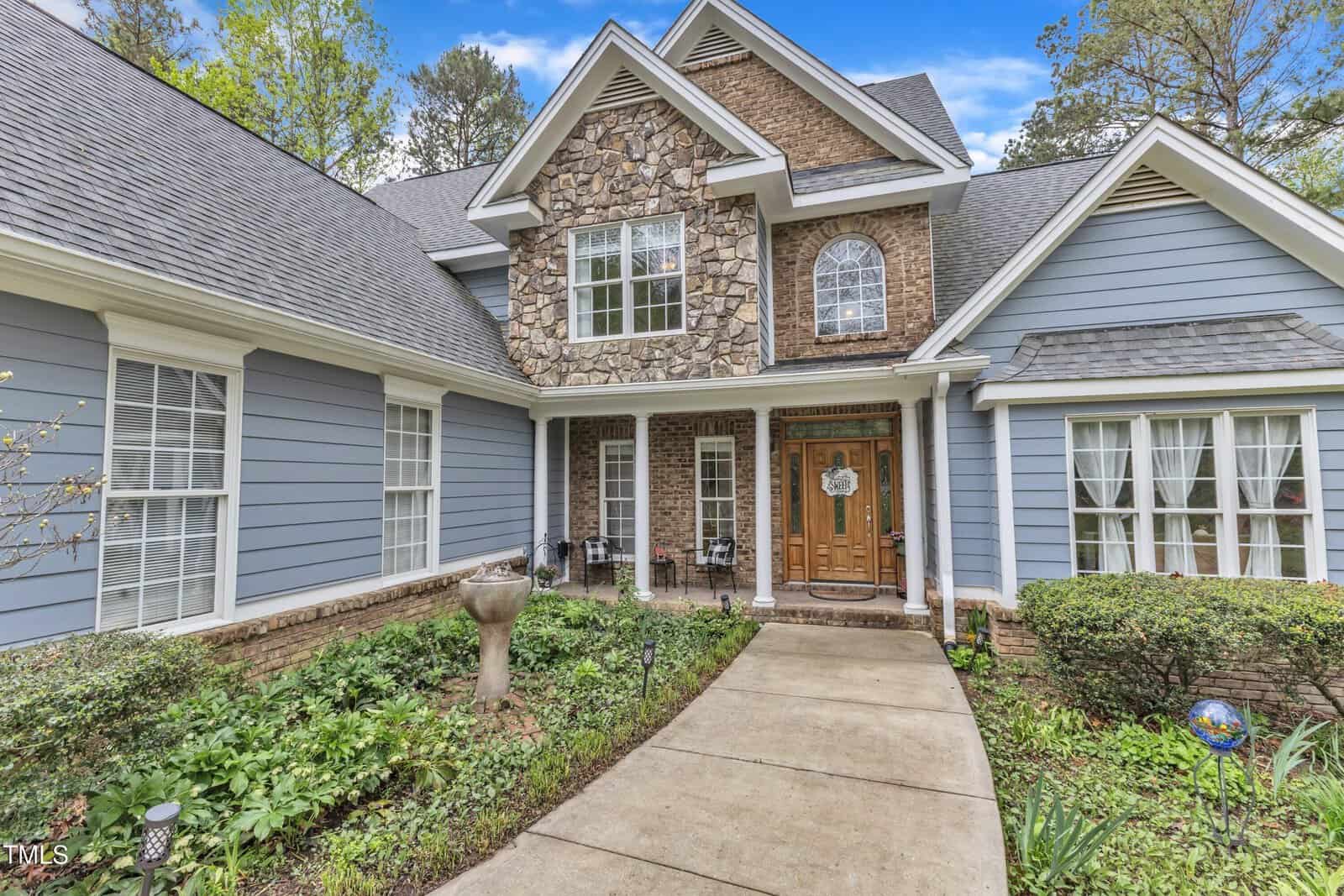 A two-story house with blue siding and a stone facade features a wooden front door with a wreath. It has a sloped roof and multiple windows. The entrance path is bordered by a landscaped garden with bushes and greenery.
