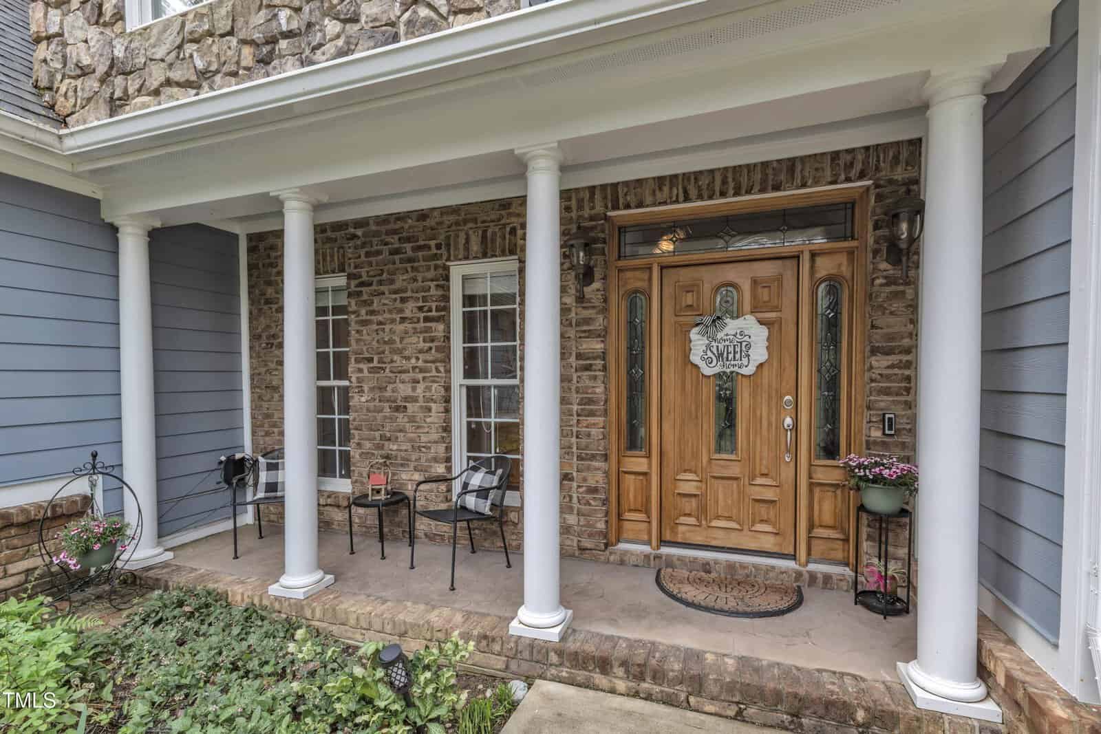 Front porch of a house with brick walls and white columns. There are two black chairs, a small table with flower pots, and a wooden door with glass panels. A sign on the door reads "Welcome Sweet Home." Steps lead up from the garden.