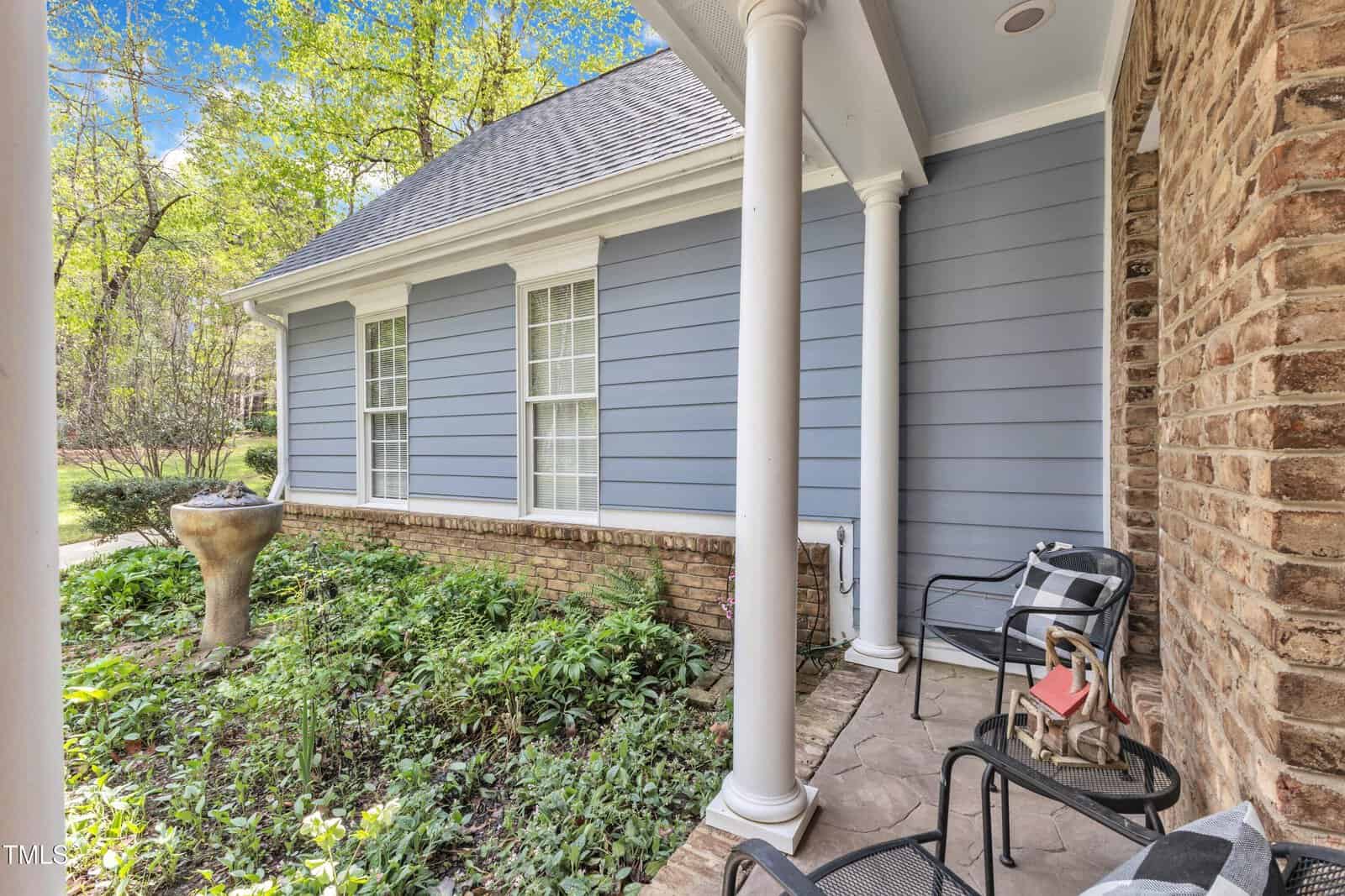 A porch with stone flooring, featuring two black metal chairs with plaid cushions and a small red table. There's a tall garden planter surrounded by plants. The house has blue siding, white windows, and brick accents. Trees are visible in the background.